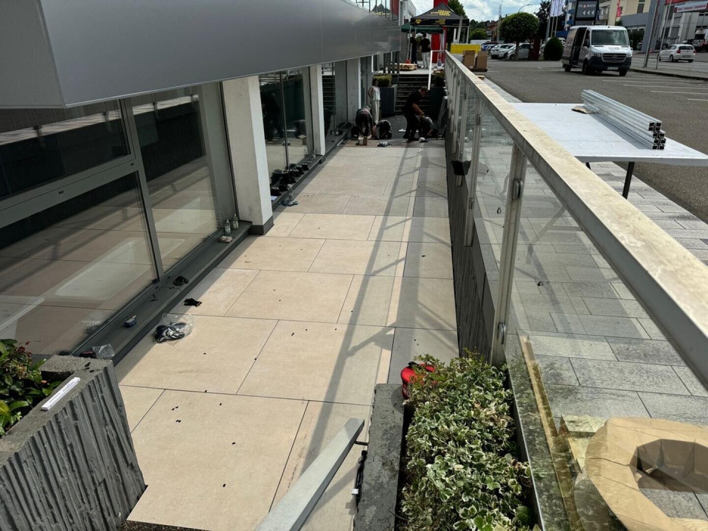 Two people work on tiling a modern outdoor terrace next to a glass railing. The area is partially tiled with large beige tiles, and tools and materials are scattered. A planter with greenery is in the foreground.