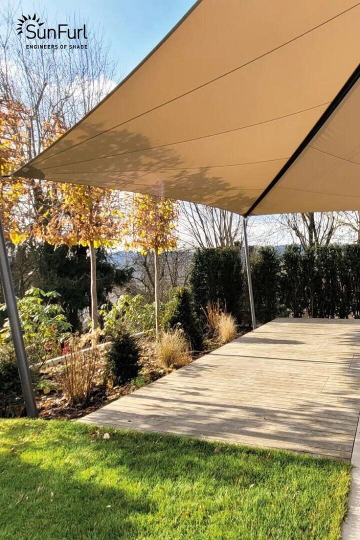 Ein großes beigefarbenes Sonnensegel ist über eine von Grün umgebene Holzterrasse gespannt, mit Bäumen und Sträuchern im Hintergrund. Das SunFurl-Logo erscheint in der oberen linken Ecke.