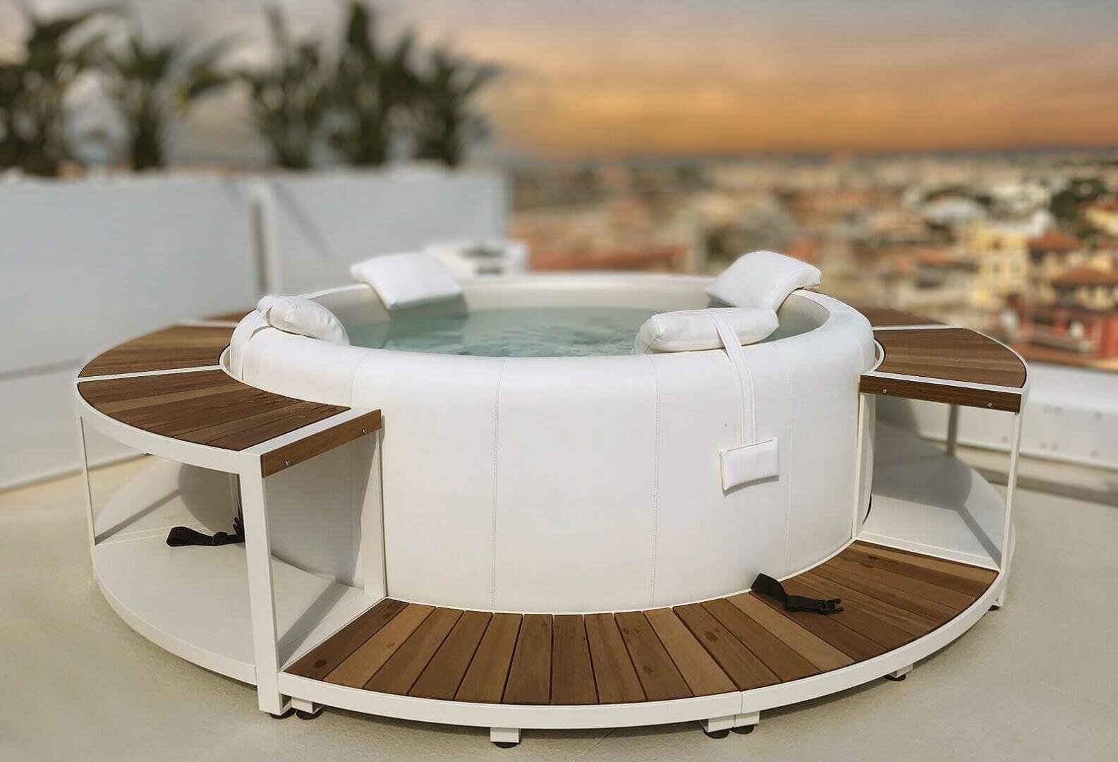 A white, round whirlpool with wooden seats and backrests stands on a roof terrace. The cityscape blurs in the background and potted plants can be seen at the edge.