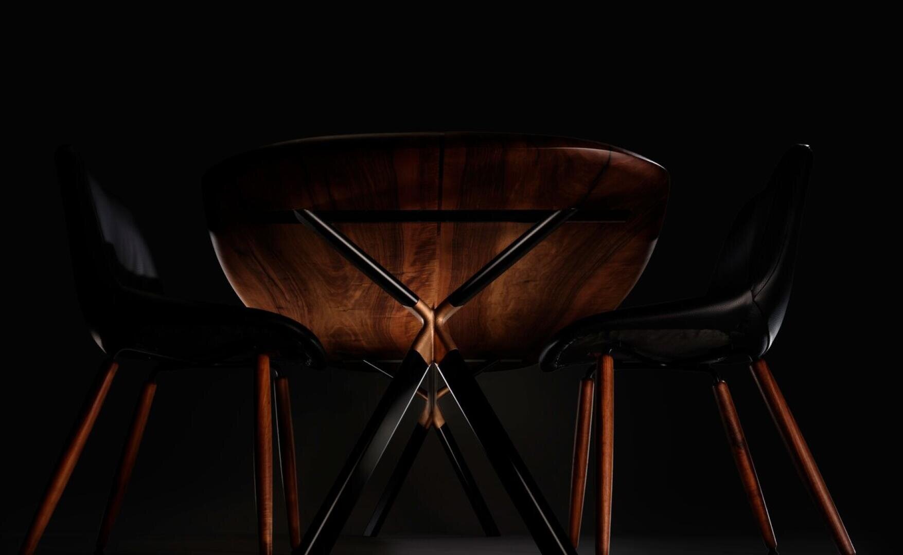A wooden table with dark legs is set against a black background, flanked by two black chairs with wooden legs. The lighting highlights the tables smooth finish and the chairs sleek design.