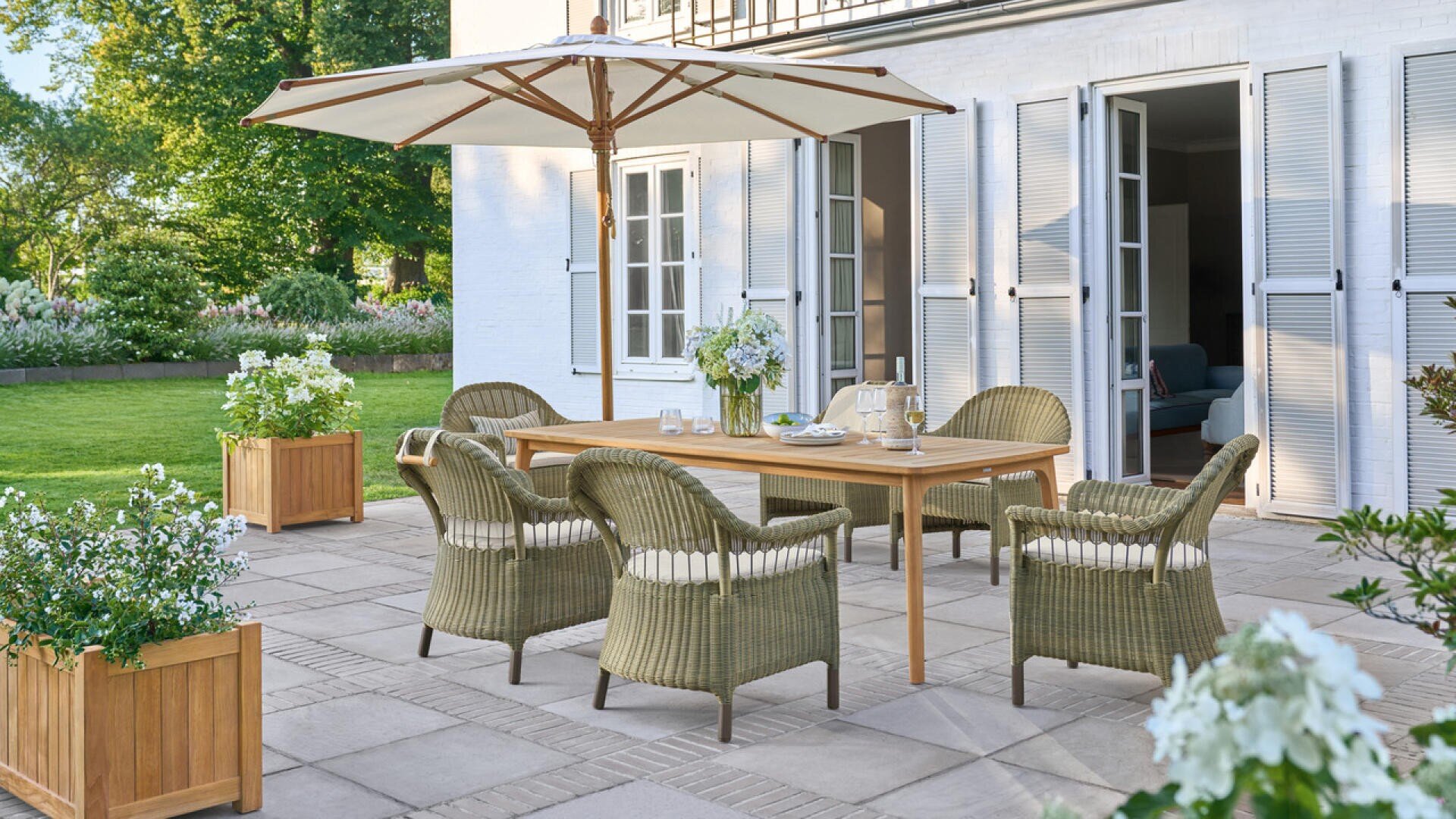 A terrace with a wooden table, five wicker chairs, a large white parasol and potted flowers, standing on a sunny day in front of a white house with open double doors and surrounded by greenery.