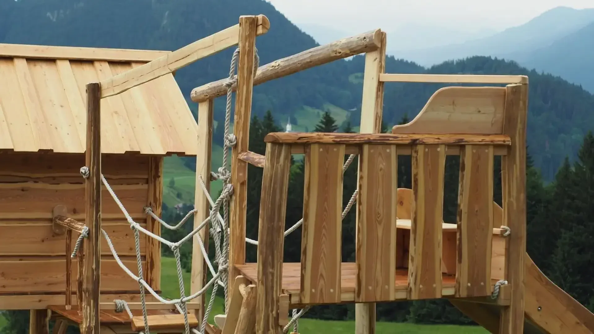 A wooden treehouse with rope railings is set on a hillside, overlooking green fields and forested mountains in the distance. The structure includes a small cabin and an open platform.