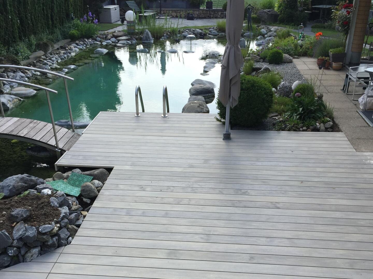 A wooden terrace leads to a natural swimming pond with clear water, surrounded by stones, green plants and patio furniture. Access is possible via a metal ladder and a small bridge leads over part of the pond.