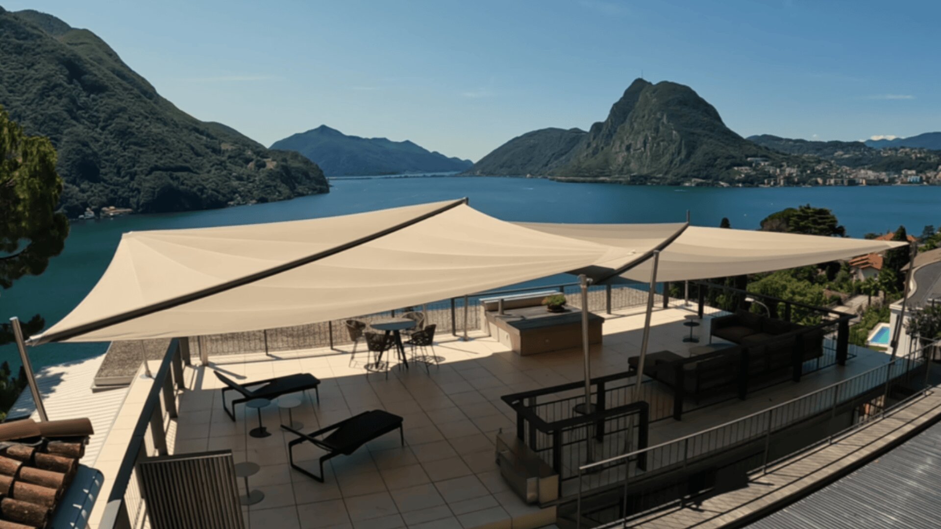 Eine geräumige Terrasse mit Liegestühlen, Tischen und großen Sonnenschirmen bietet einen Blick auf einen ruhigen blauen See, der von grünen Bergen umgeben ist, und einen klaren Himmel.