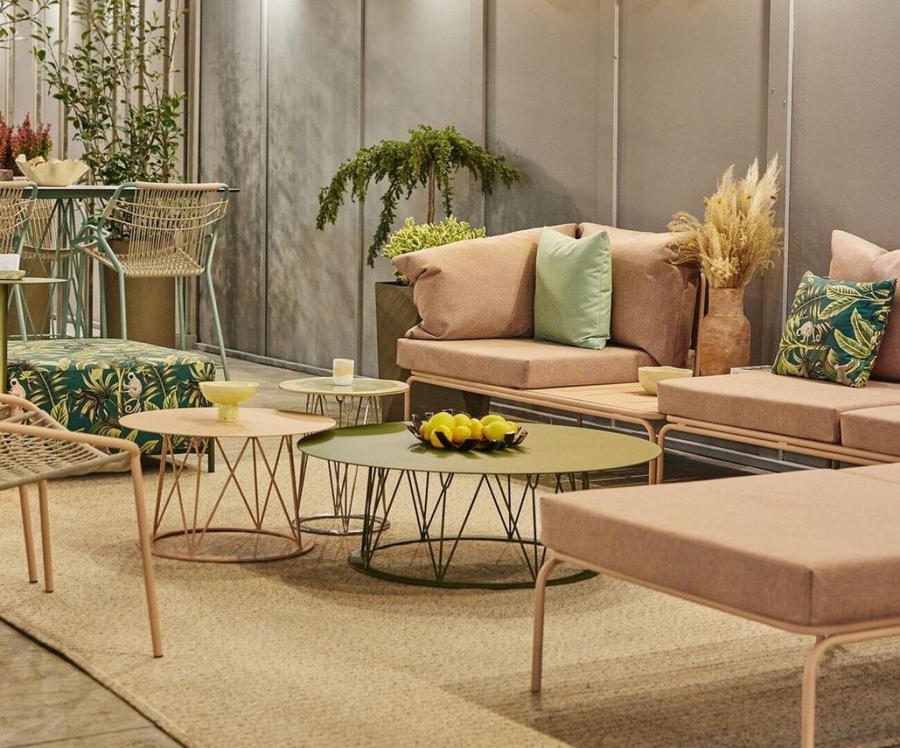 Moderner Sitzbereich im Freien mit beigefarbenen gepolsterten Sofas, grünen Kissen, runden Couchtischen mit Drahtgestell, einem Mittelstück aus Zitronen und dekorativen Pflanzen, alles auf einem gewebten Teppich vor einer grauen Wand.