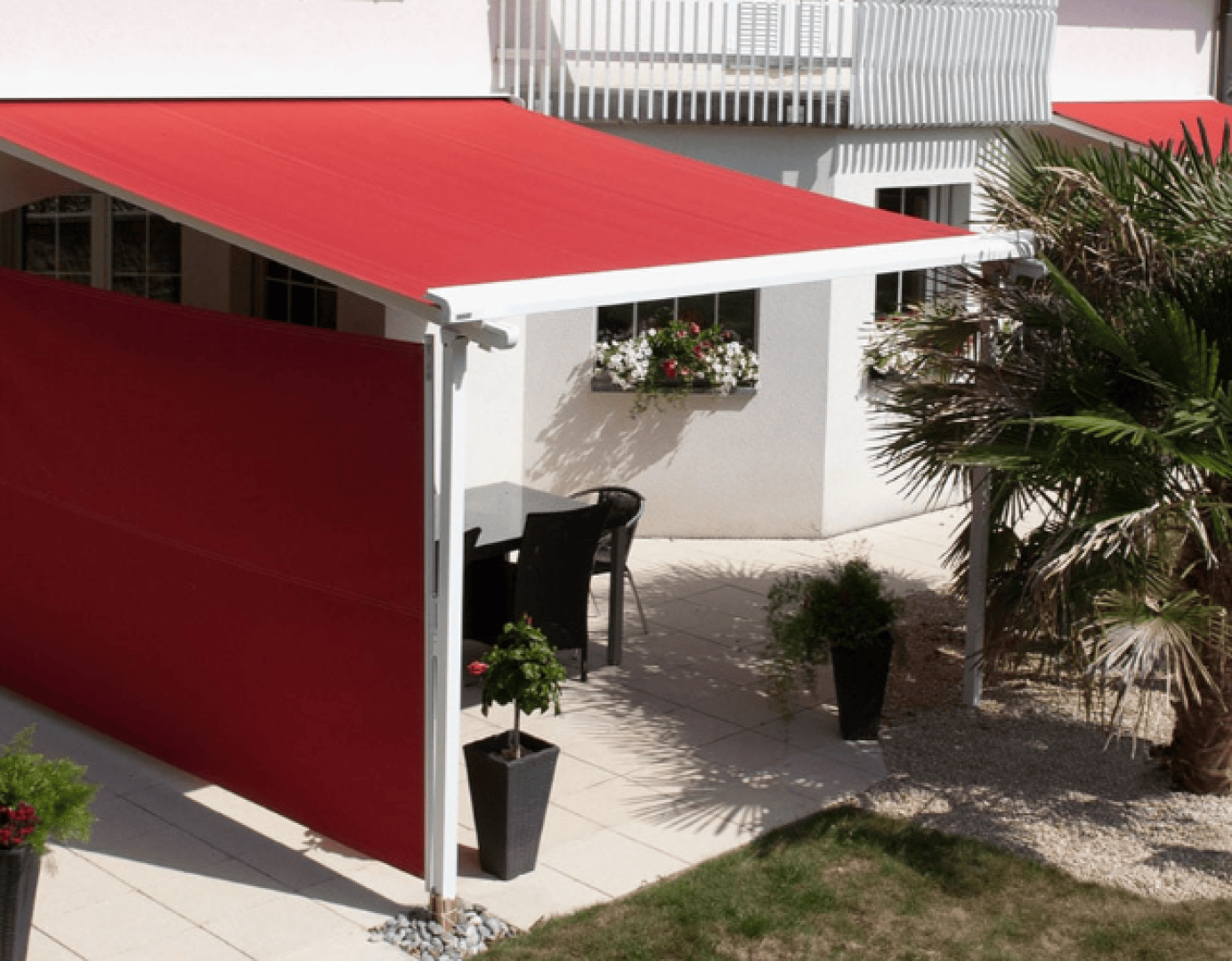A patio with a red awning and matching red side shade, outdoor table and chairs, potted plants, and a palm tree, adjacent to a white building with window flower boxes.