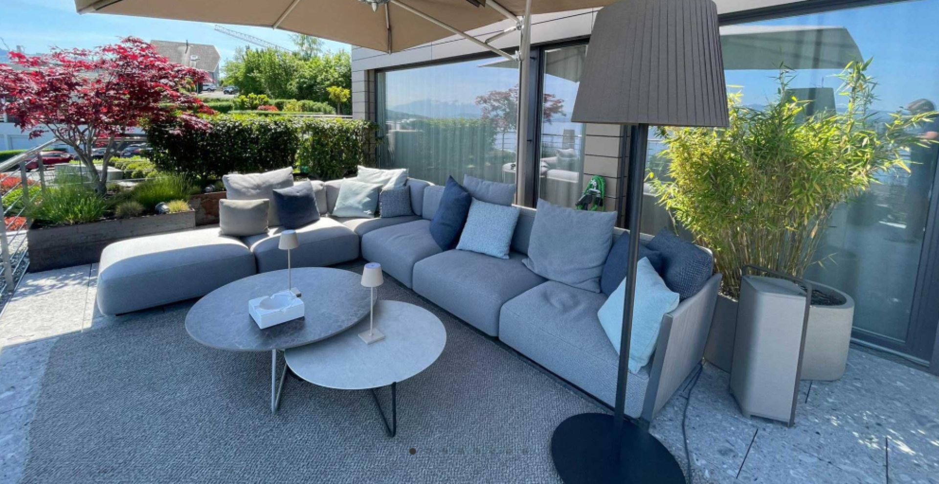 Moderne Außenterrasse mit einem großen grauen Sektionssofa, blauen und grauen Polstern, runden Couchtischen, mehreren Lampen und Topfpflanzen. Ein großer Sonnenschirm spendet Schatten und im Hintergrund befindet sich ein üppiger Garten.