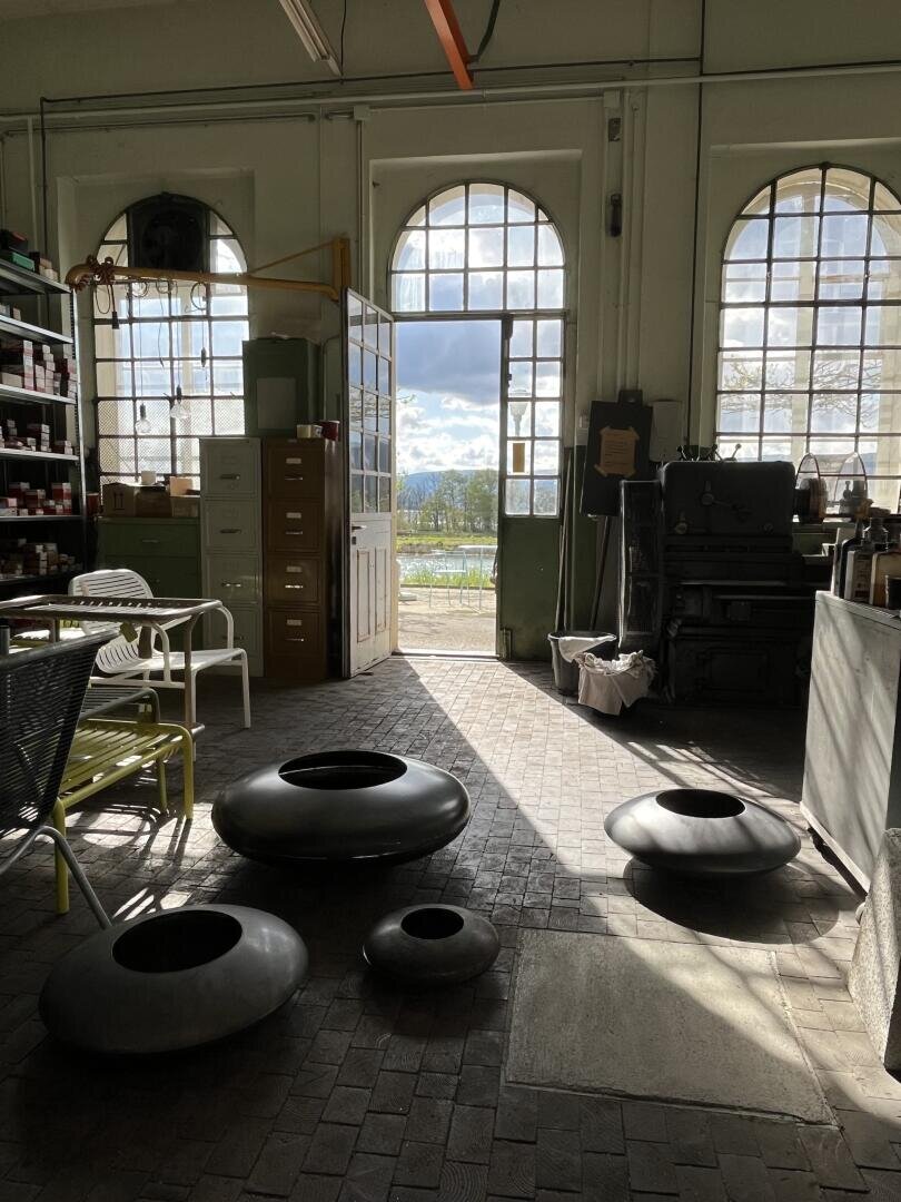 A sunlit workshop with large arched windows, metal shelves, filing cabinets, and round black sculptures on a brick floor. Outside, trees and mountains are visible through the windows.