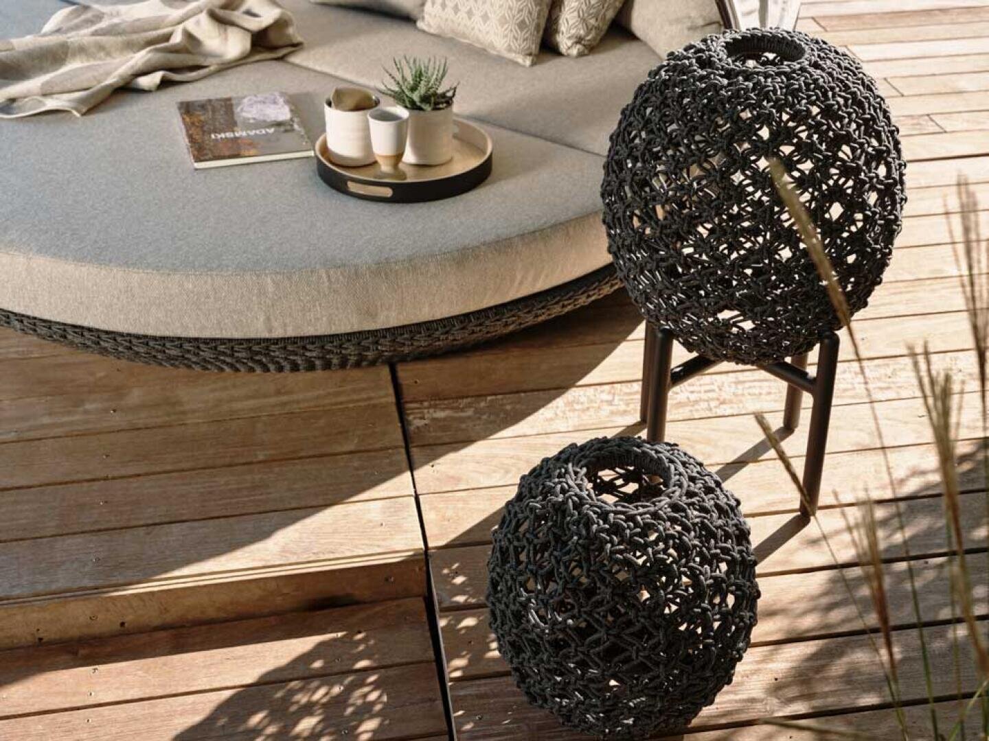 Eine gemütliche Sitzecke im Freien mit einem runden gepolsterten Sofa, einem Tablett mit Tassen und einer Pflanze sowie zwei geflochtenen dekorativen Laternen auf einer Holzterrasse. Das warme Sonnenlicht wirft weiche Schatten auf die Szene.