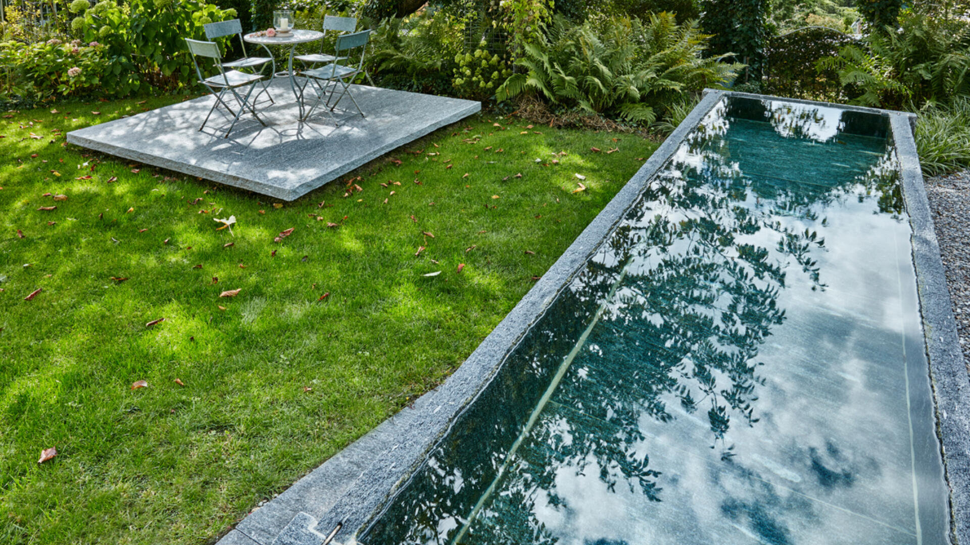Ein schmaler, rechteckiger, spiegelnder Pool neben einer Rasenfläche mit verstreutem Laub; im Hintergrund befindet sich eine kleine Steinterrasse mit einem Bistrotisch und zwei Stühlen unter belaubten Bäumen.