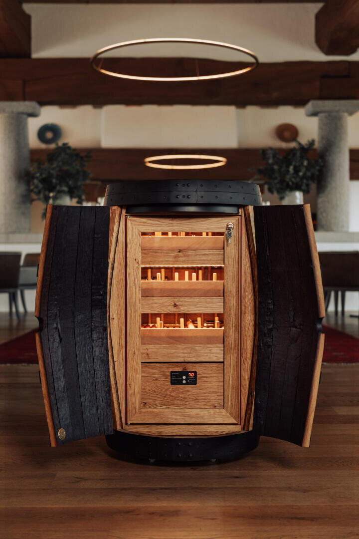 A wooden barrel converted into a humidor stands in a modern room whose doors open to reveal orderly shelves of cigars, softly illuminated by the interior lighting.