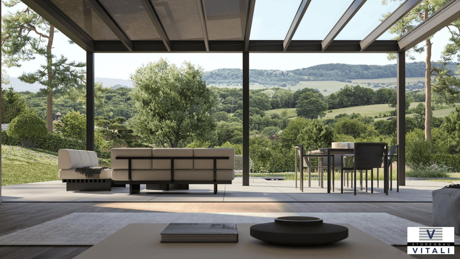 Ein moderner, offener Wohnbereich mit raumhohen Glaswänden bietet einen Blick auf eine üppige grüne Landschaft und ferne Hügel. Auf der Terrasse sind Sitzgelegenheiten und Essmöbel zu sehen. In der Ecke befindet sich ein Logo mit der Aufschrift Stoneware Vitali.