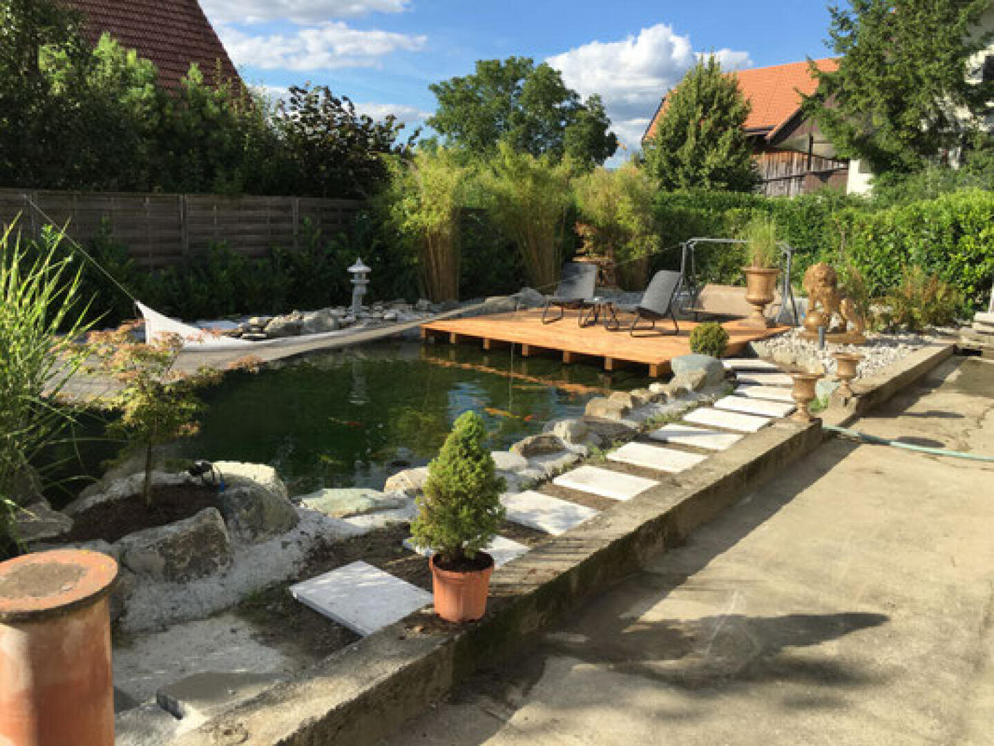 A backyard pond with koi fish, surrounded by rocks and greenery, features a wooden deck with lounge chairs and a hammock. Potted plants and garden statues decorate the peaceful outdoor space.