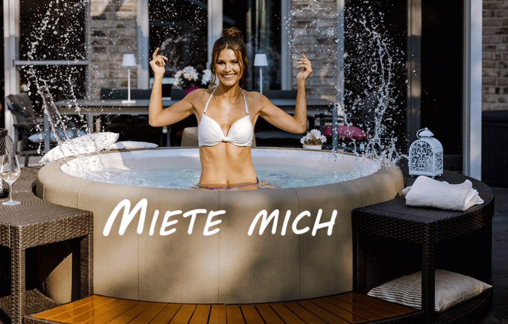 A smiling woman in a white bikini splashes water while standing in an outdoor inflatable hot tub. The words Miete mich (German for Rent me) are written across the tub. Outdoor furniture and flowers are nearby.