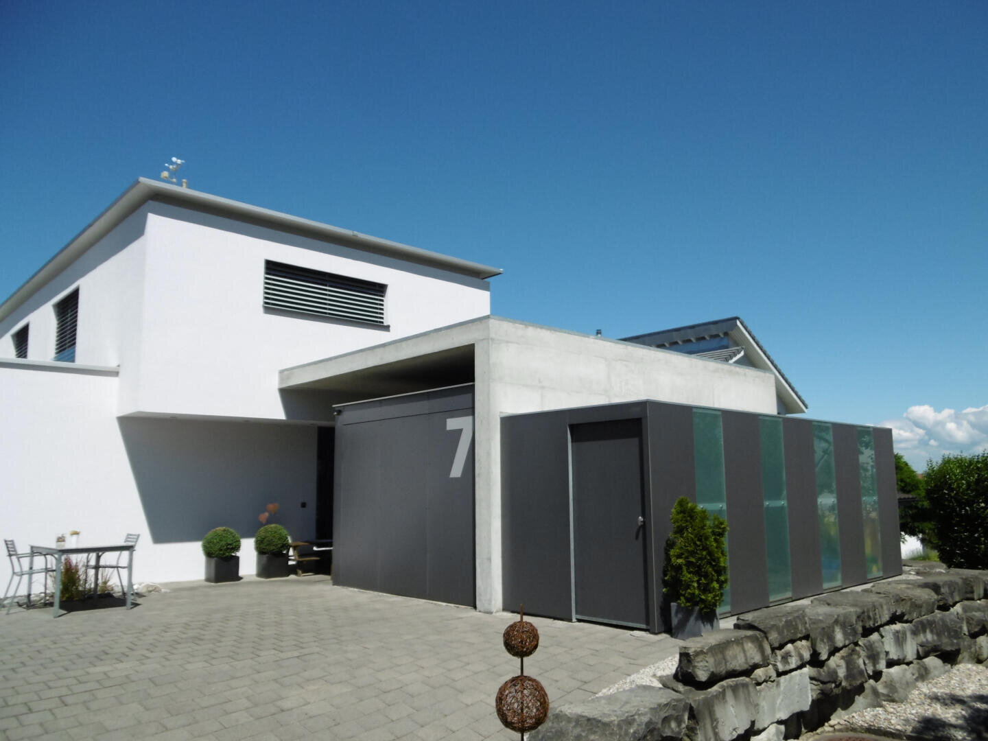 Ein modernes weißes Haus mit flachen Dächern, großen grauen Garagentoren mit einer "7", kleinen Sträuchern in Pflanzkübeln und einer gepflasterten Einfahrt unter einem klaren blauen Himmel.
