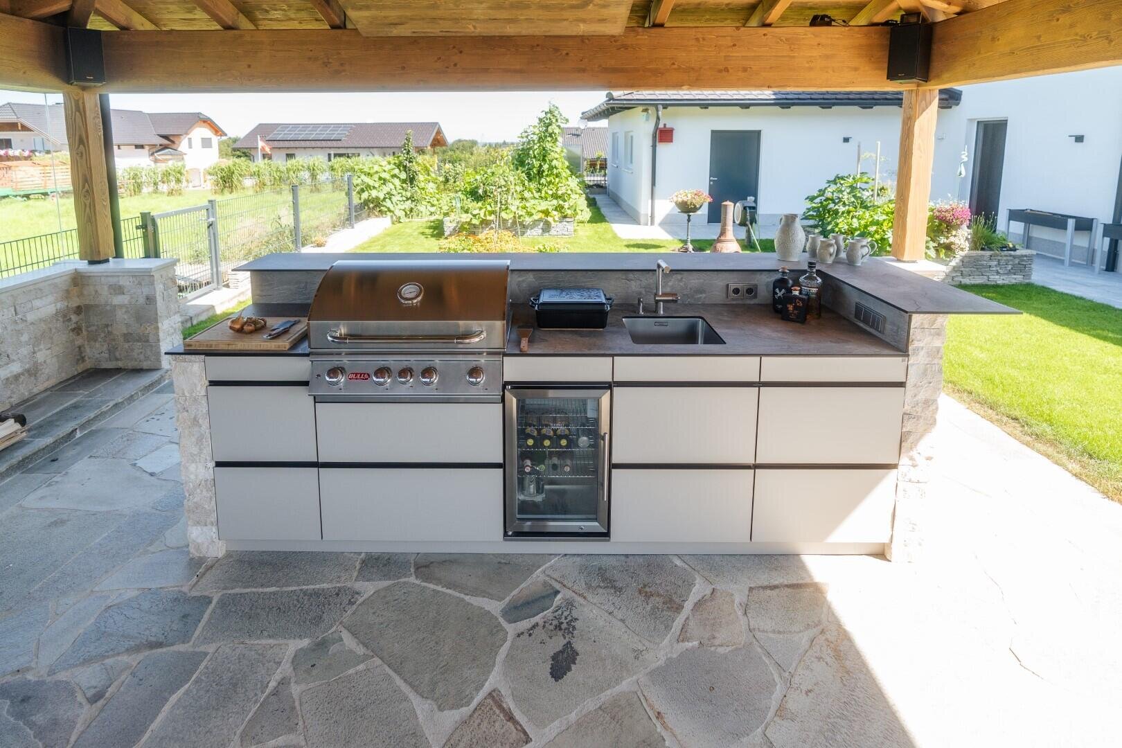 Moderne Außenküche unter einer hölzernen Pergola mit eingebautem Grill, Spüle, Kühlschrank, Arbeitsplatte und Schränken auf einer Steinterrasse mit Blick auf einen Garten und die umliegenden Häuser.