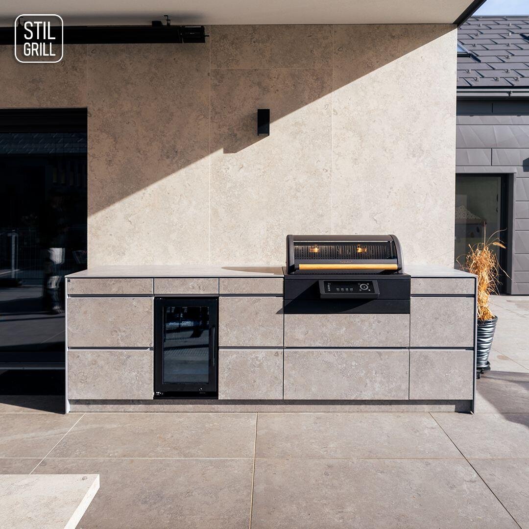 Moderne Außenküche mit hellgrauen Steinschränken, einem eingebauten schwarzen Grill, einem kleinen Getränkekühlschrank und minimalistischem Design; auf einer Terrasse mit neutralem Bodenbelag gelegen. STIL GRILL-Logo in der oberen linken Ecke.
