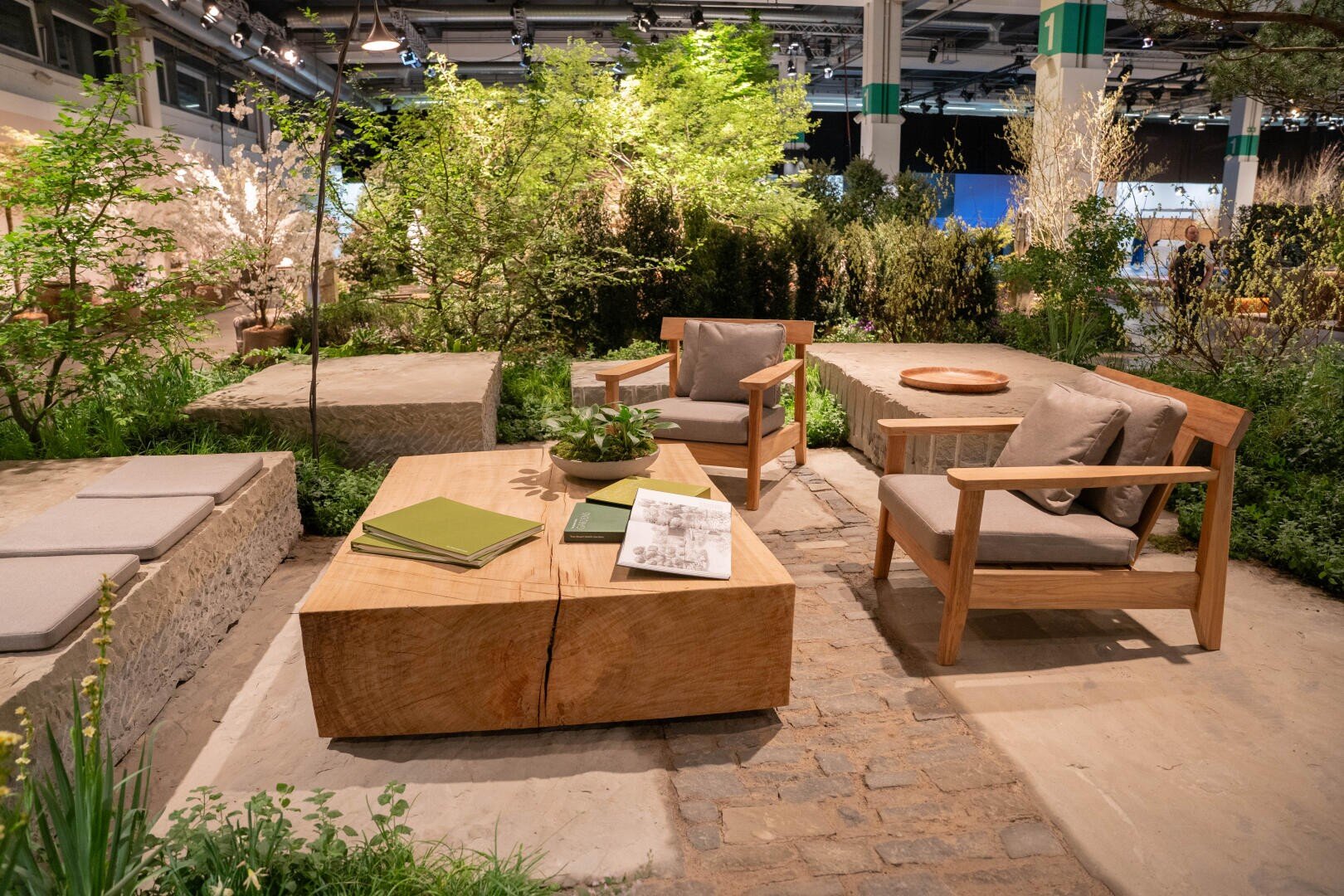 Ein moderner Sitzbereich im Freien mit Holzstühlen, Steinbänken und einem hölzernen Couchtisch, umgeben von üppigen grünen Pflanzen und Bäumen, auf einem gepflasterten Boden im Innenbereich mit heller Beleuchtung.
