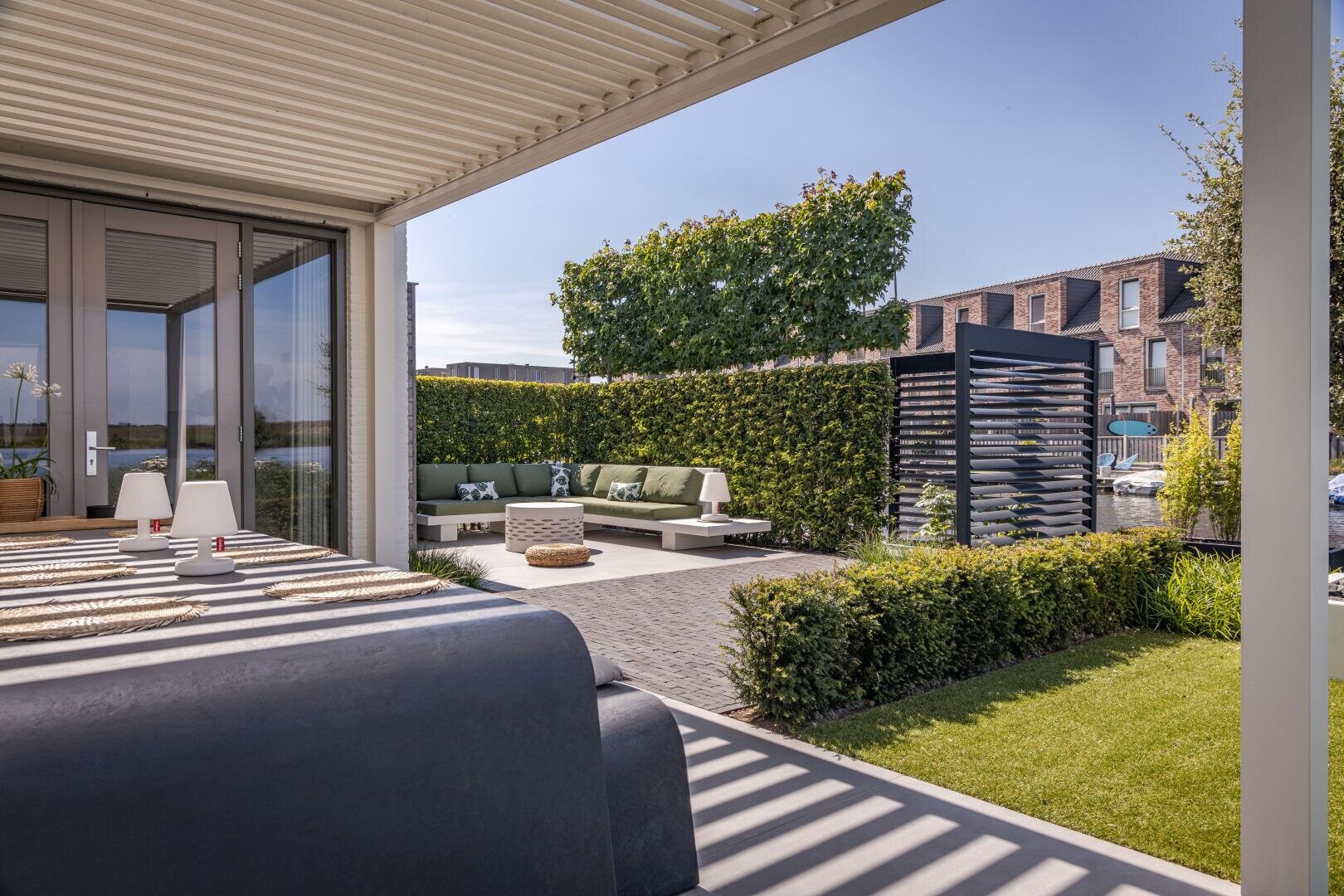 Modern outdoor terrace with a covered dining area, a cushioned seating area in the corner, neatly trimmed hedges, a decorative privacy screen and modern houses in the background under a clear blue sky.