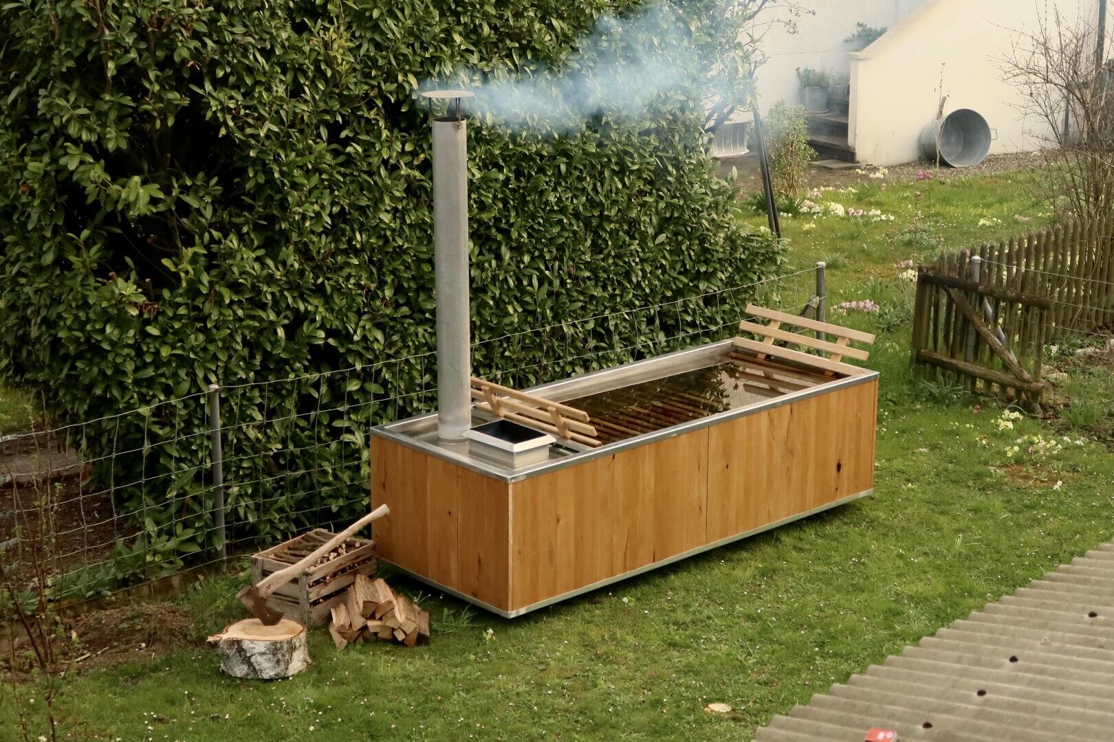 A wooden hot tub with a metal chimney smokes in a grassy yard. Firewood and an axe are stacked nearby, and two wooden benches stand next to and on top of the tub. A fence and green plants can be seen in the background.