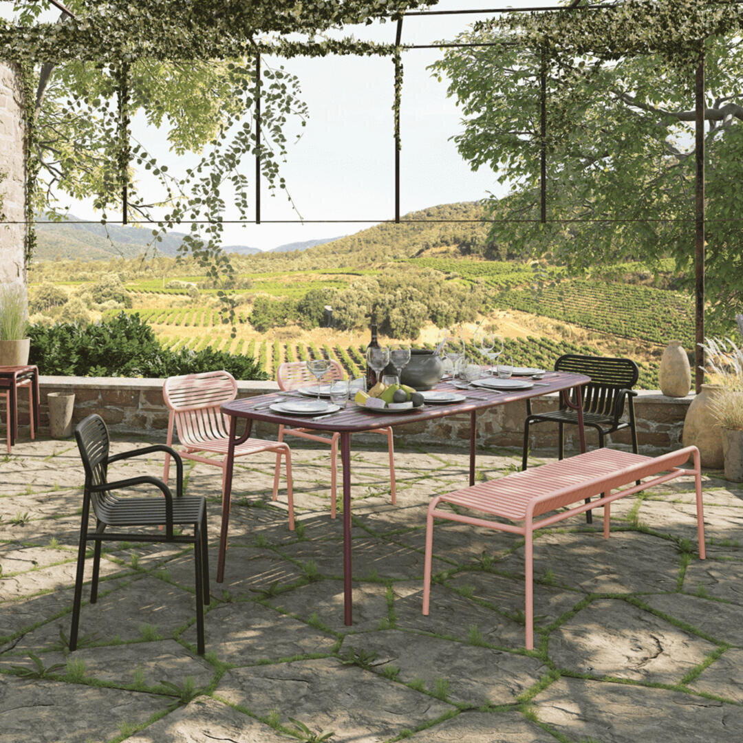 Outdoor dining area with a table set for a meal, surrounded by modern chairs and benches on a stone patio. Lush vineyards and rolling hills are visible in the background, under a pergola with creeping vines.