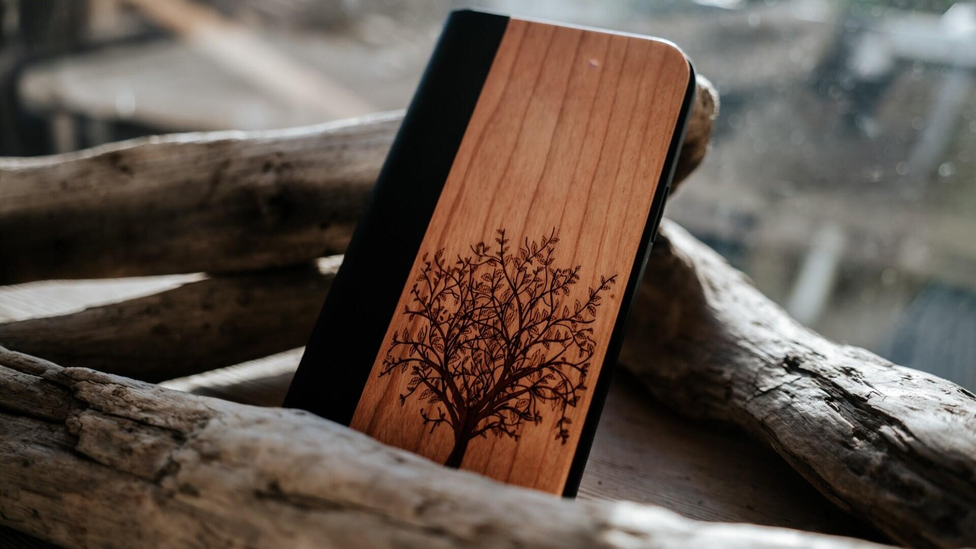 Ein hölzernes Notizbuch mit schwarzem Buchrücken und einem eingravierten Baummuster auf dem Einband liegt auf einem Stück Treibholz. Das natürliche Licht hebt die Texturen des Notizbuchs und des Holzes hervor.