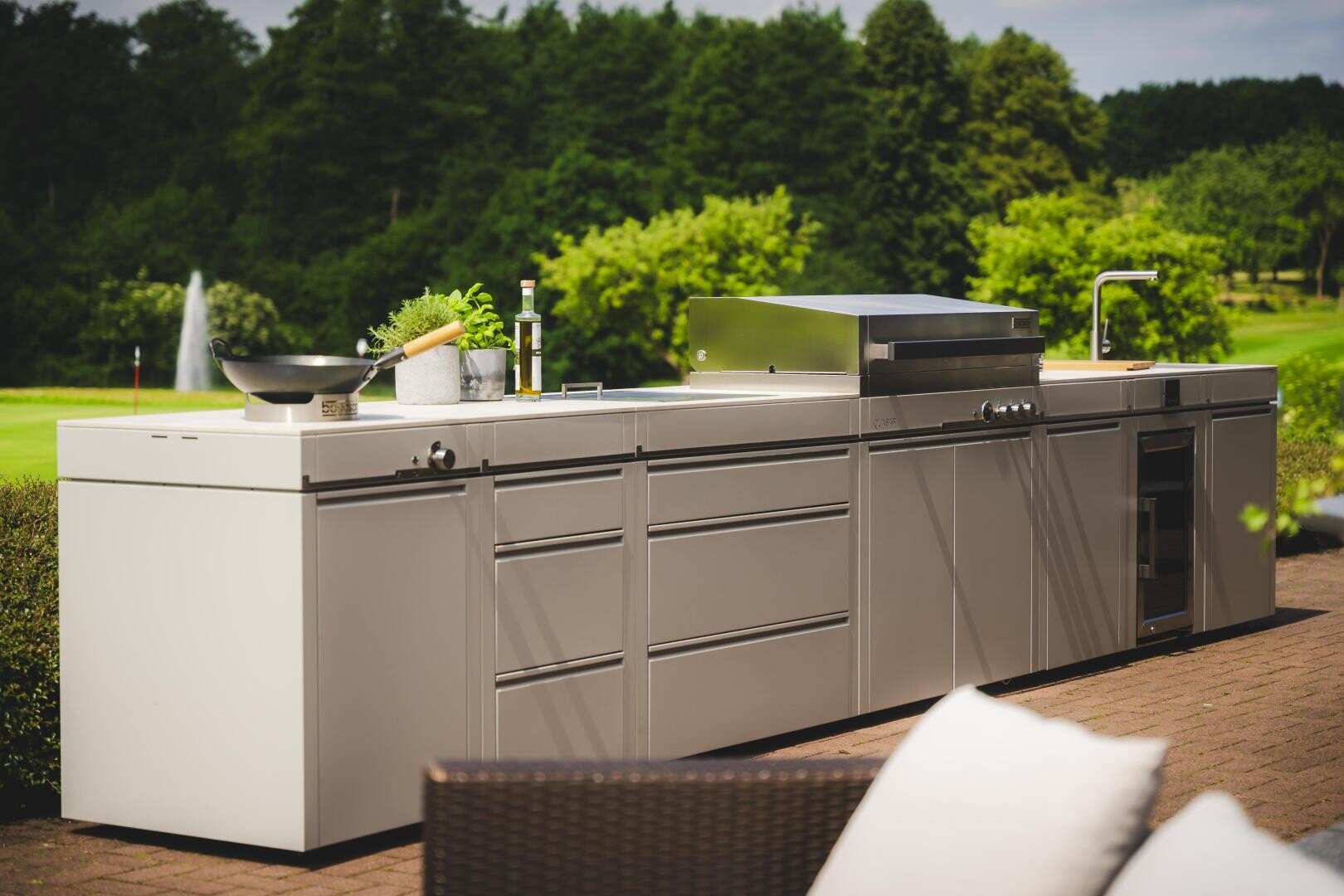 Eine moderne Außenküche mit Schränken aus rostfreiem Stahl, einem eingebauten Grill, einem Spülbecken und Kochutensilien auf einer Terrasse mit grünen Bäumen und einem Springbrunnen im Hintergrund.