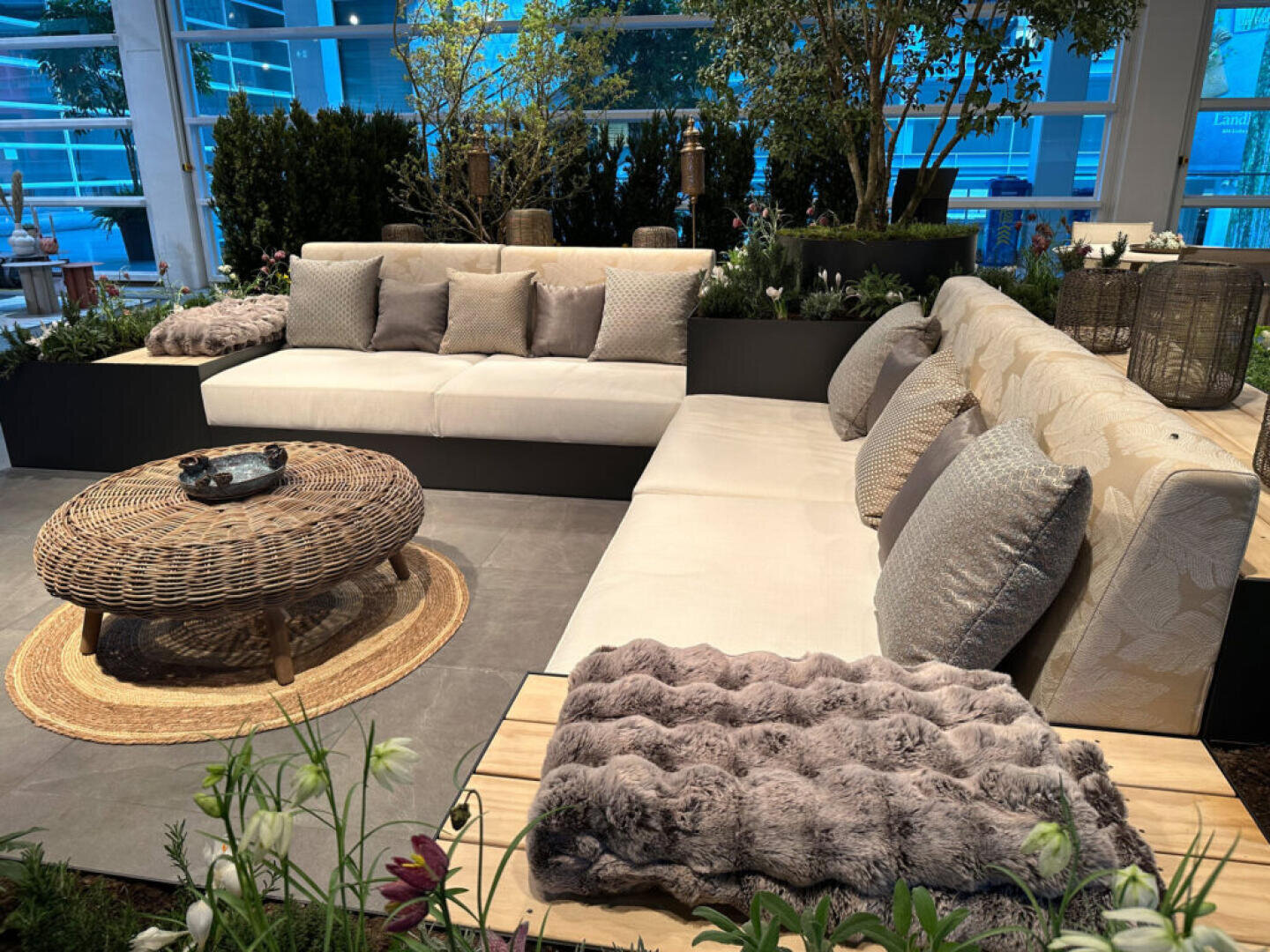 Moderner Sitzbereich im Freien mit zwei cremefarbenen Sofas, grauen und beigen Kissen, einem runden Couchtisch aus Korbgeflecht auf einem geflochtenen Teppich, üppigen Grünpflanzen und natürlichem Dekor in einem hellen, verglasten Raum.