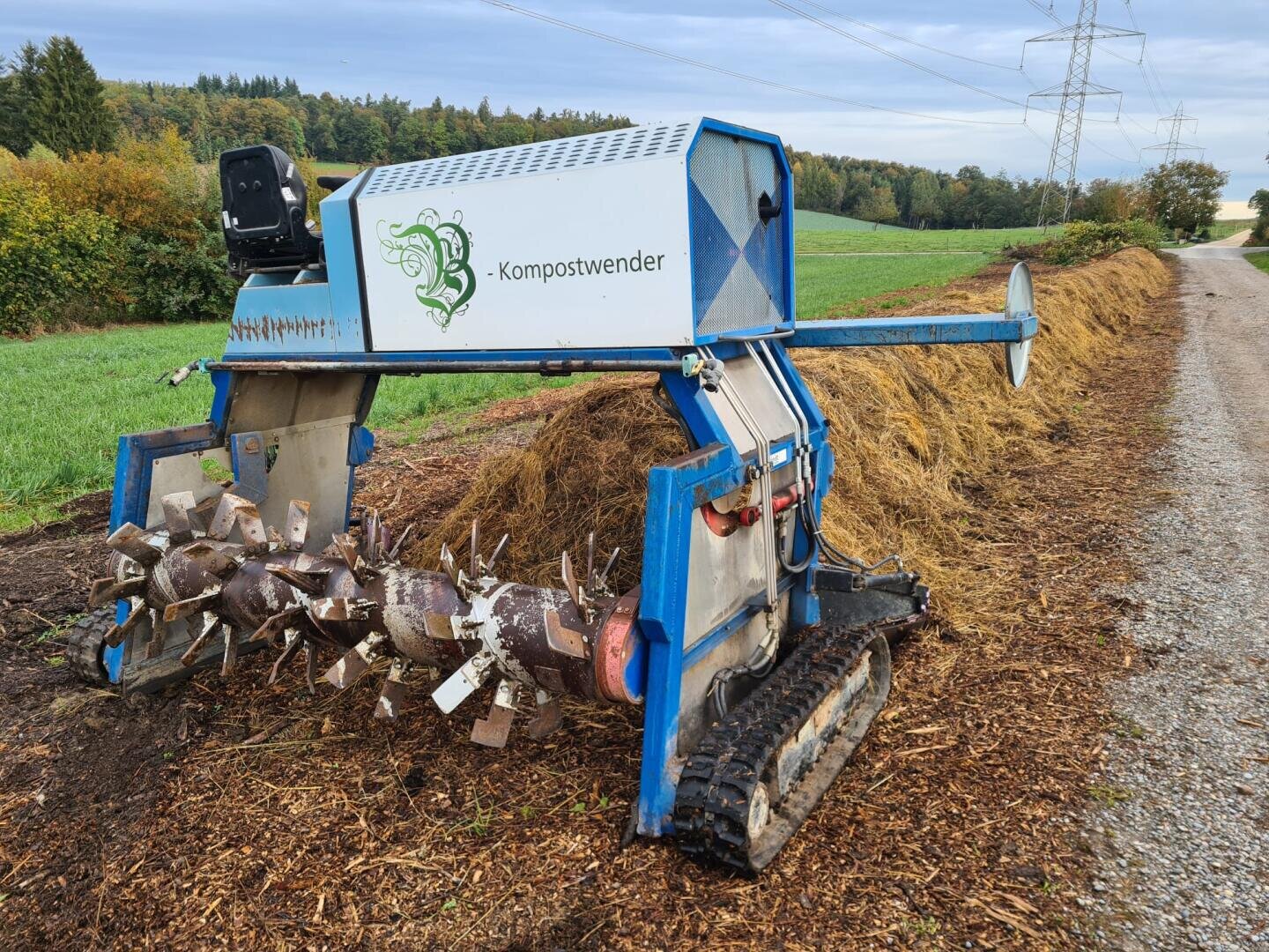 A blue and silver compost turner stands on a dirt road next to a long pile of compost and mulch, with green fields and trees under a cloudy sky in the background.