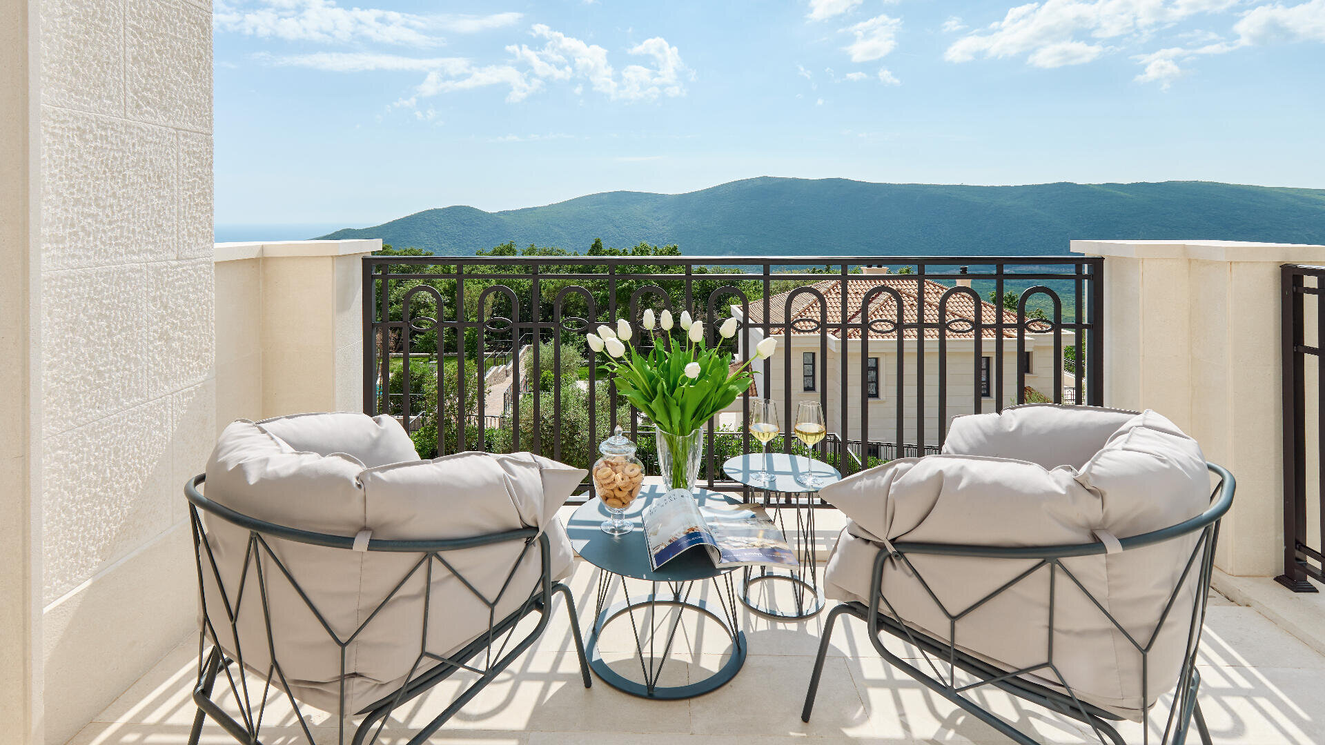 Ein heller Balkon mit zwei gepolsterten Stühlen, einem kleinen Tisch mit Getränken, Snacks und Blumen bietet einen Blick auf grüne Hügel und das ferne Meer unter einem teilweise bewölkten Himmel.
