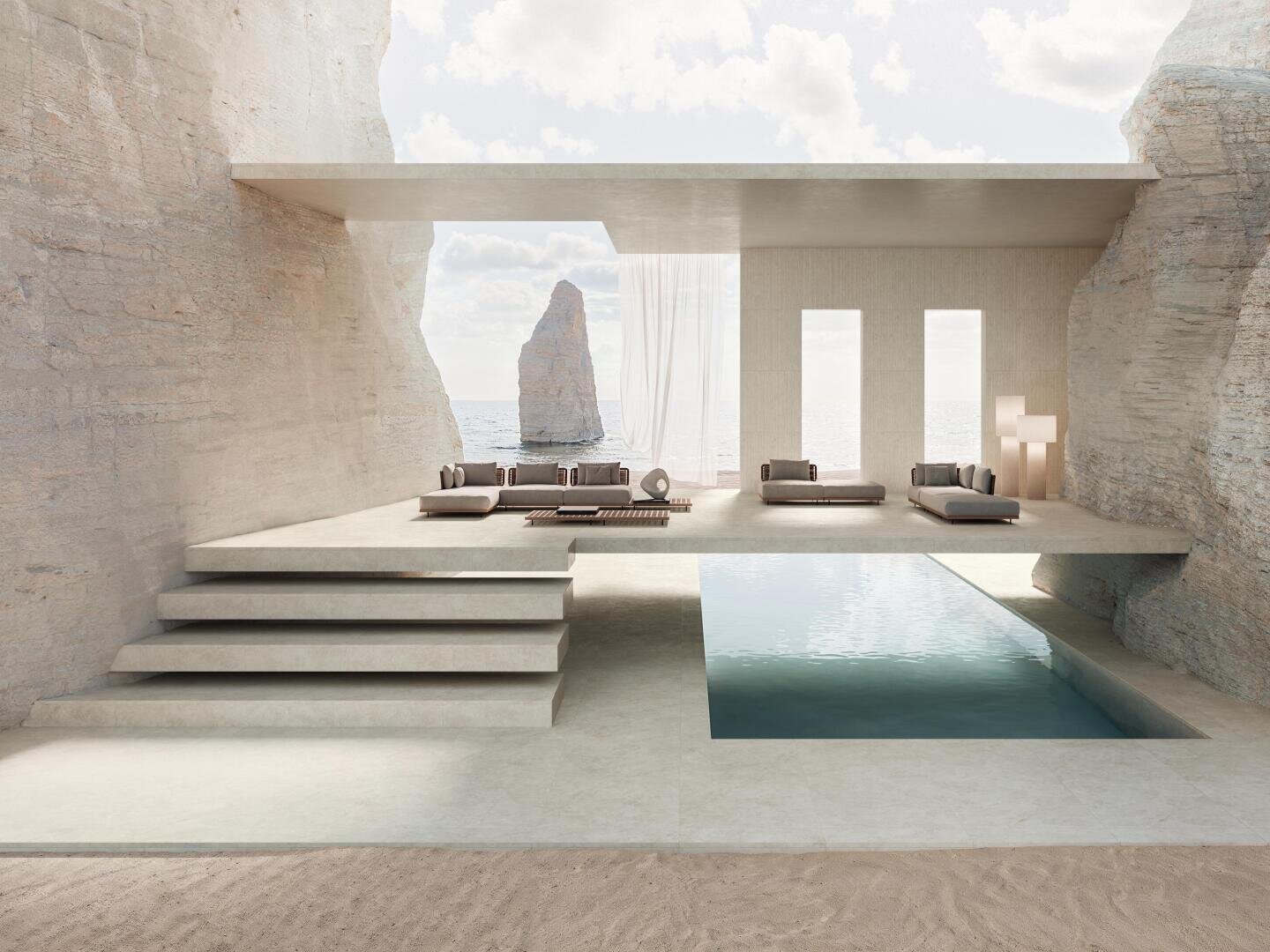 Minimalistischer, in die Felsen gehauener Open-Air-Wohnbereich mit rechteckigem Pool, modernen Sofas, durchsichtigen Vorhängen und Blick auf den Ozean mit einer hohen Felsformation in der Ferne unter einem teilweise bewölkten Himmel.