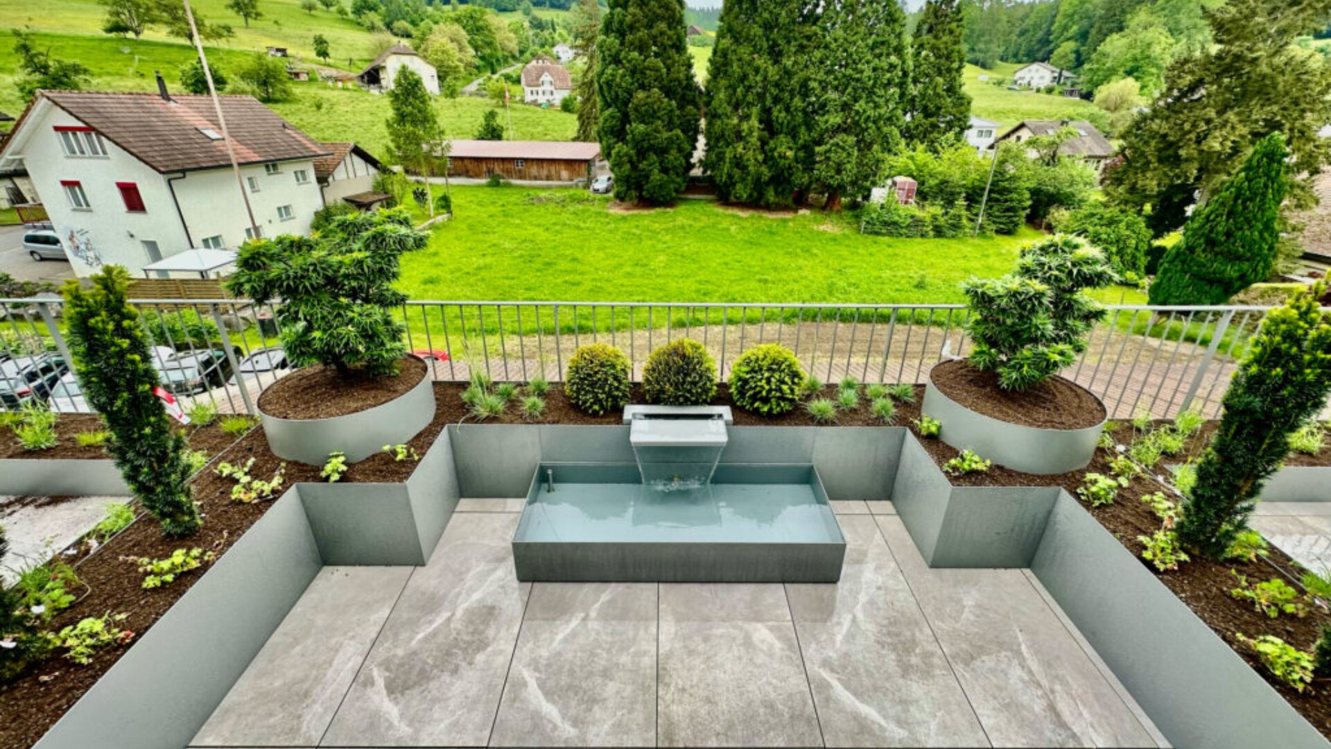 Ein moderner Dachgarten mit einem kleinen rechteckigen Wasserbrunnen, ordentlich angeordneten Pflanzgefäßen mit Sträuchern und Bäumen, mit Blick auf grüne Felder, Häuser und hohe Bäume im Hintergrund.