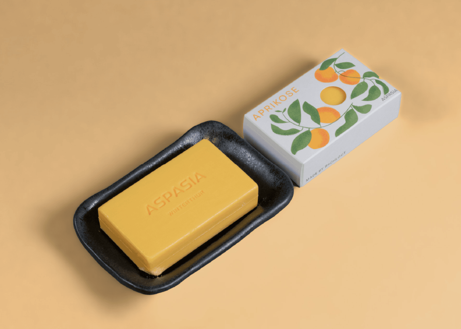 A yellow bar of soap with the inscription ASPASIA NATURKOSMETIK rests on a black dish next to the box, which is decorated with illustrations of apricots and labeled APRIKOSE.