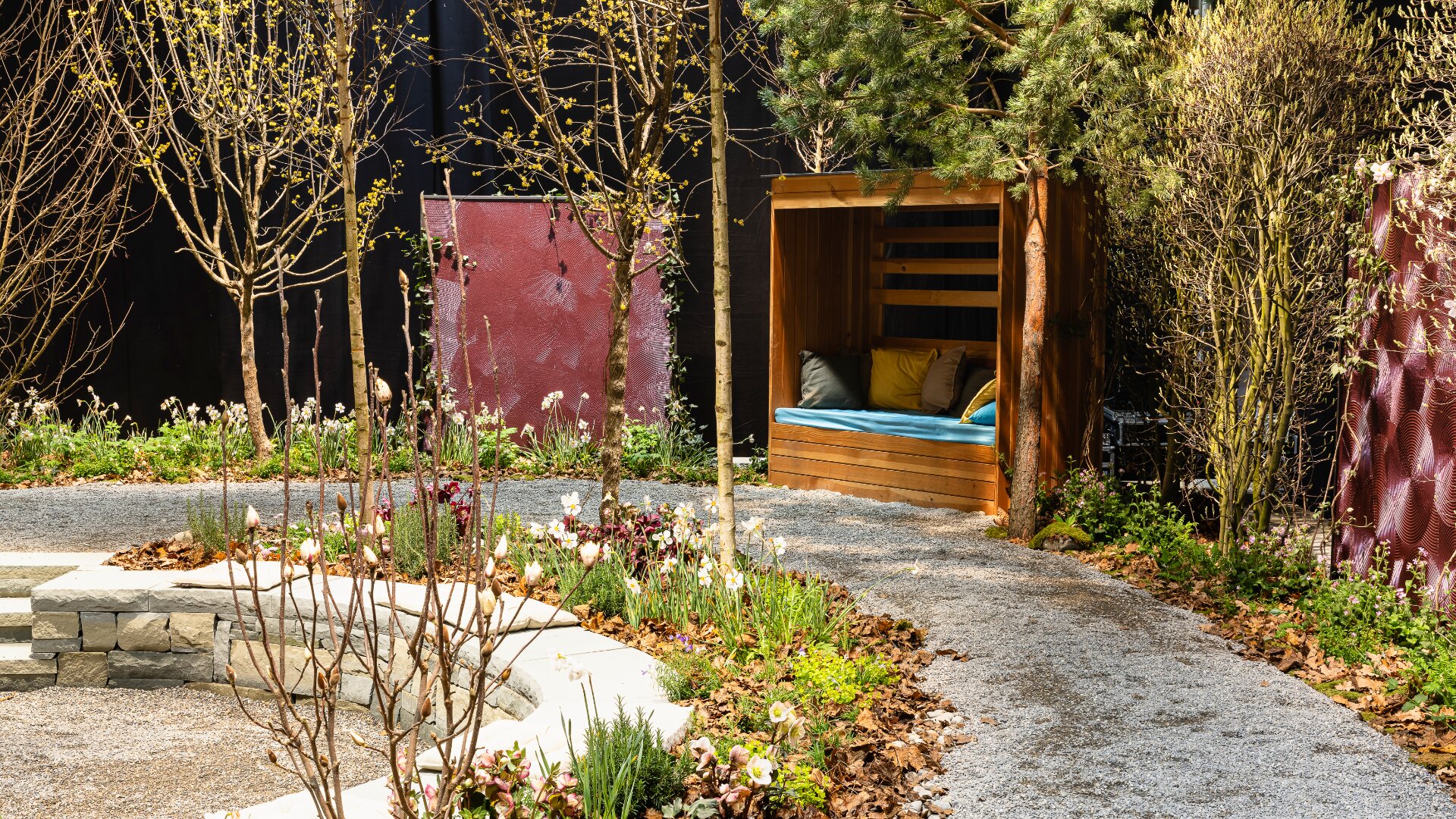 Ein angelegter Garten mit einem gewundenen Kiesweg, Blumenbeeten und jungen Bäumen. In der Ecke befindet sich eine hölzerne Sitzecke mit blauen und gelben Kissen, umgeben von Grünpflanzen und lila Sichtschutzwänden.