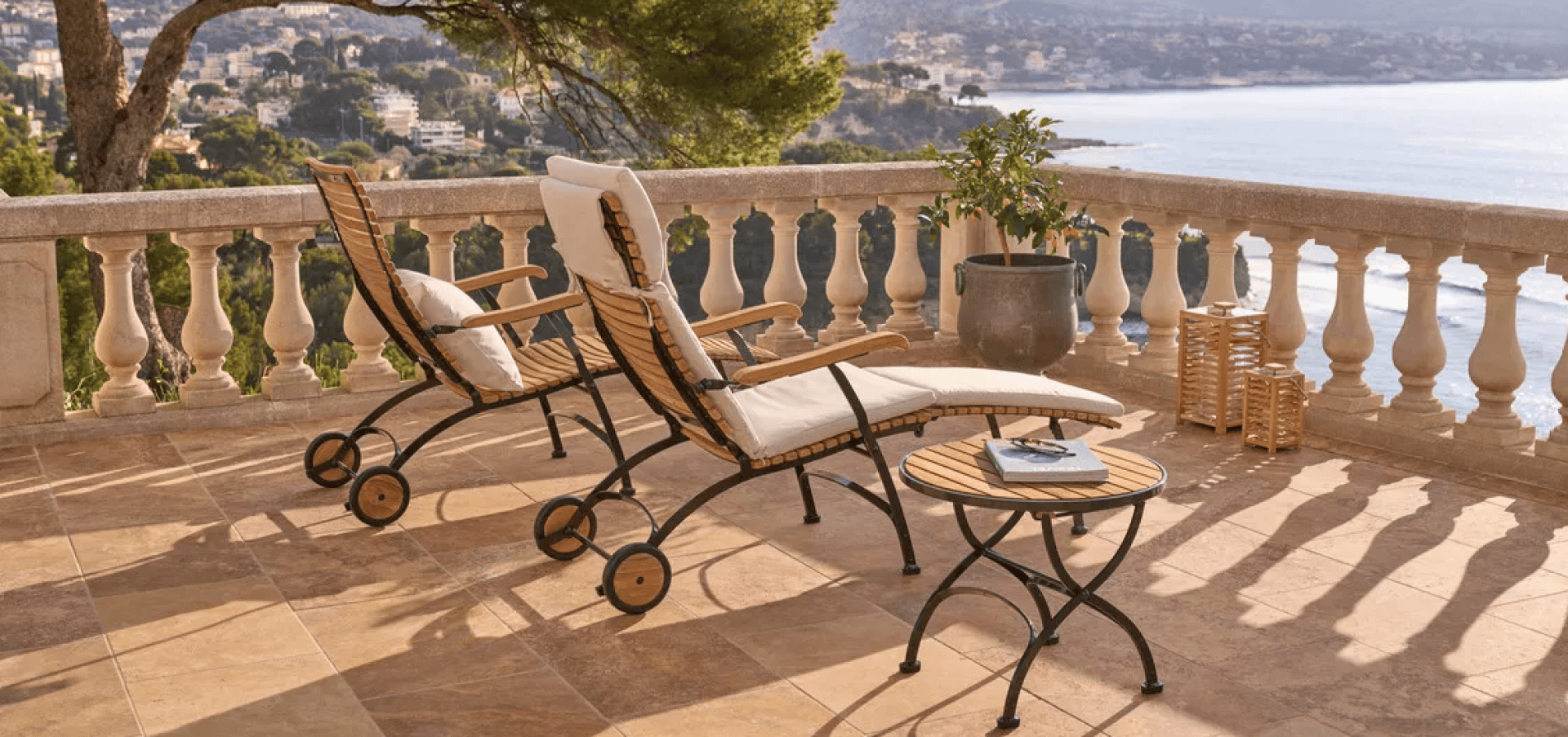 Two cushioned lounge chairs and a small round table sit on a sunlit stone terrace with a scenic view of the sea, hills, and distant houses. A potted plant and wicker basket are nearby.