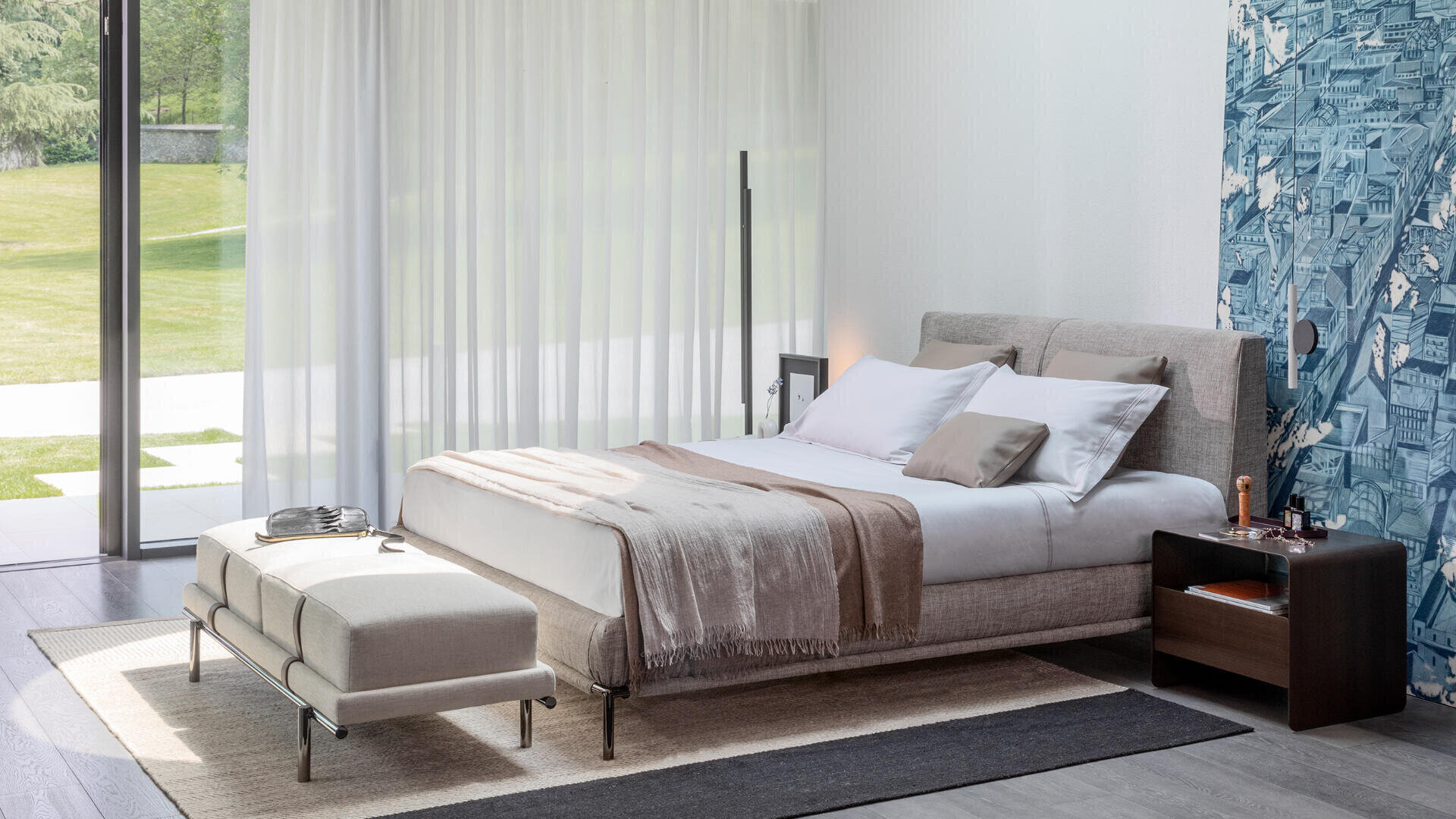 A modern bedroom with a large bed, beige bed linen and a neutral-colored blanket. There is a bench at the foot of the bed. The room has floor-to-ceiling windows with white curtains and a patterned blue accent wall.