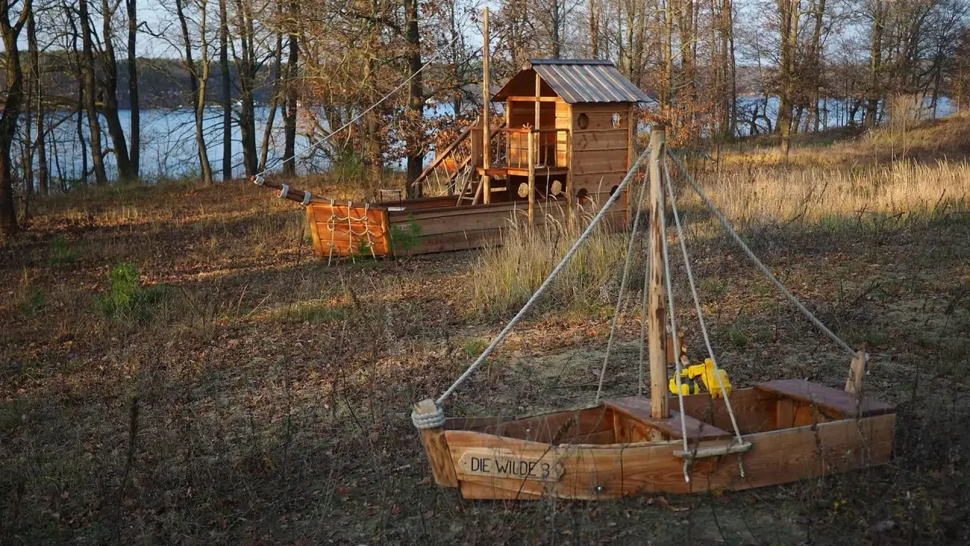 Zwei kleine hölzerne Spielboote und ein Spielhaus liegen auf einer grasbewachsenen, bewaldeten Fläche in der Nähe eines Sees. Die Boote und das Haus sind aus Brettern gebaut, und an einem sonnigen Tag ist das Gebiet von blattlosen Bäumen umgeben.