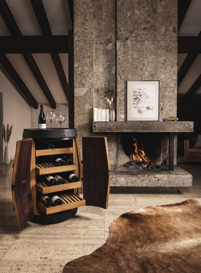 A wine barrel cabinet with open doors displays wine bottles and glasses in front of a stone fireplace with a burning fire, framed artwork above and a cowhide rug on the floor in a cozy living room.