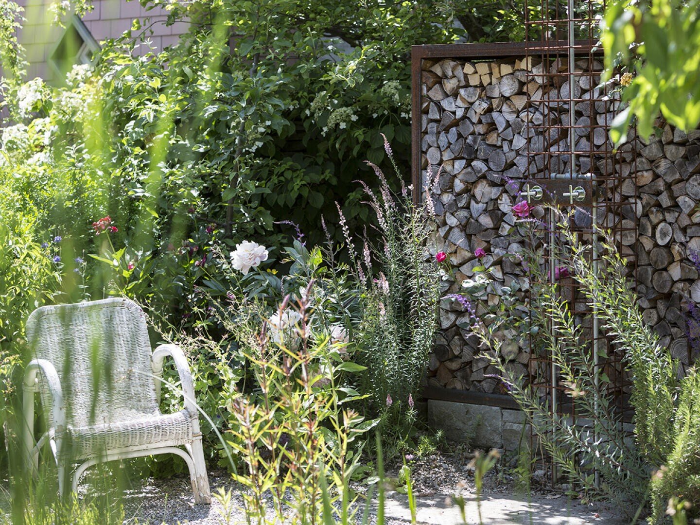 Ein sonnenbeschienener Garten mit einem weißen Korbstuhl, Wildblumen, üppigen Grünpflanzen und einer Wand mit Brennholz neben einem Metallgitter. Das gedämpfte Sonnenlicht schafft eine friedliche, einladende Atmosphäre.
