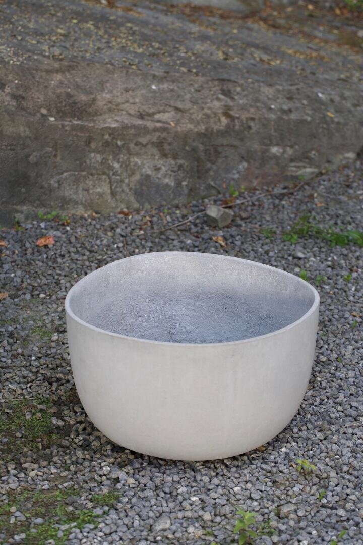 Eine große, runde, weiße Keramik- oder Betonschale steht im Freien auf Kies, mit einer Felswand und verstreutem Grün im Hintergrund.
