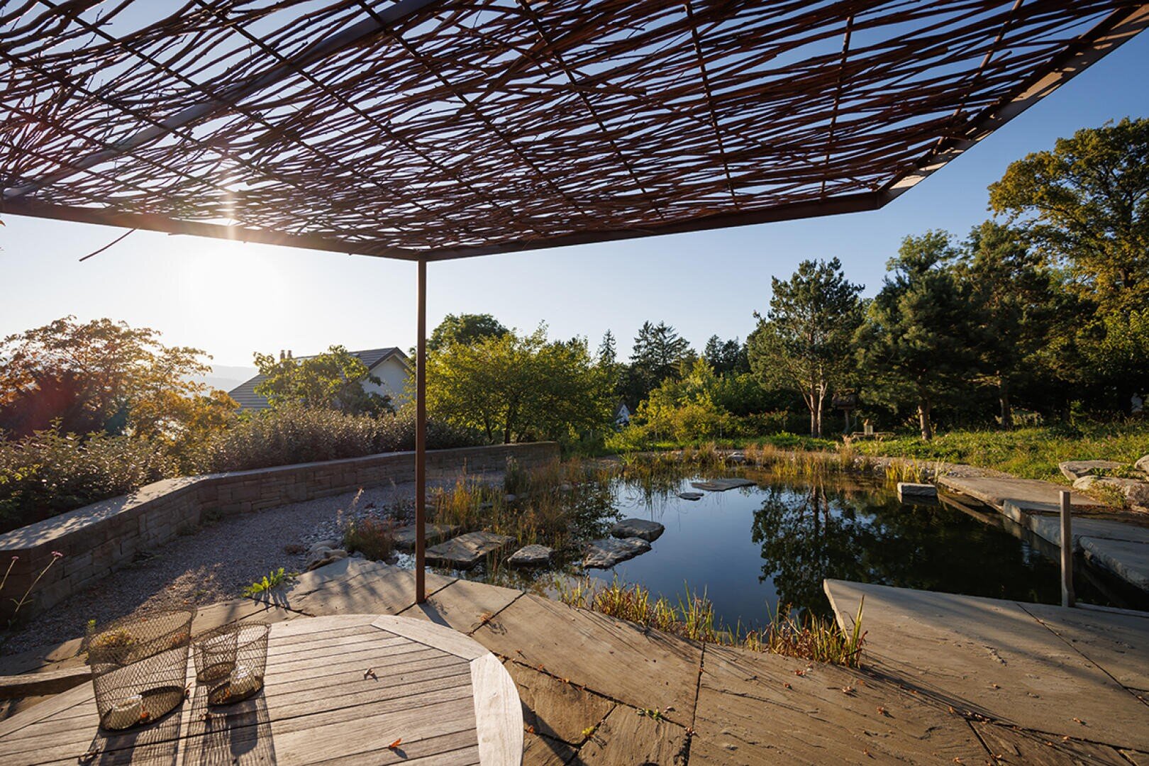 Eine moderne Gartenterrasse mit einem Holztisch, zwei Stühlen und einem Schattenspender aus geflochtenen Ästen blickt auf einen natürlichen Teich, der im späten Nachmittagssonnenlicht von Bäumen und Grün umgeben ist.
