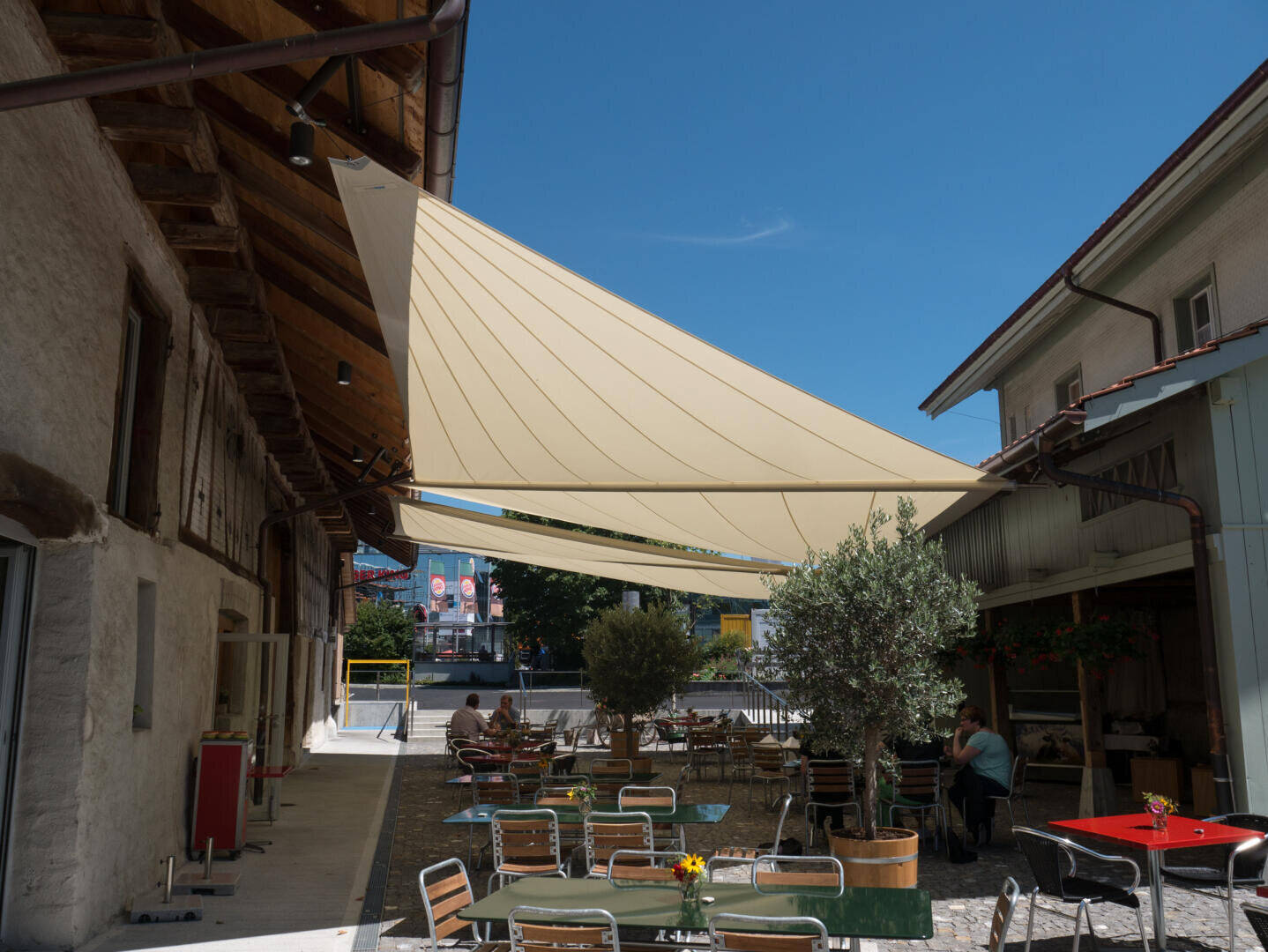 Außencafé mit Tischen und Stühlen unter großen beigen Sonnensegeln zwischen zwei Gebäuden, sonniges Wetter, blauer Himmel und einige Topfpflanzen rund um den Sitzbereich.