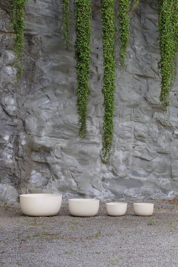 Vier cremefarbene runde Töpfe unterschiedlicher Größe sind auf einer Kiesfläche vor einer grauen Steinmauer aufgereiht, von oben hängen lange grüne Ranken herab.