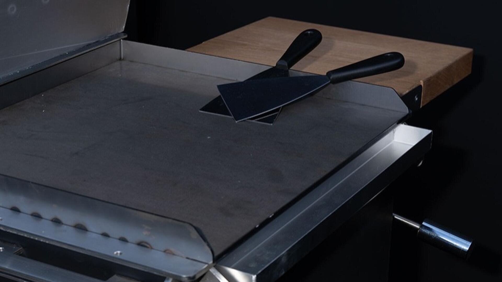 Eine Grillplatte aus Metall mit zwei gekreuzten flachen Spateln und schwarzen Griffen. Die Bratpfanne hat eine hölzerne Seitenablage und steht vor einem dunklen Hintergrund.