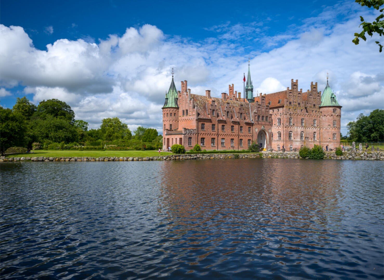 Ein großes Backsteinschloss mit grünen Dächern steht an einem ruhigen See unter einem blauen Himmel mit vereinzelten Wolken, umgeben von üppig grünen Bäumen und Gras.
