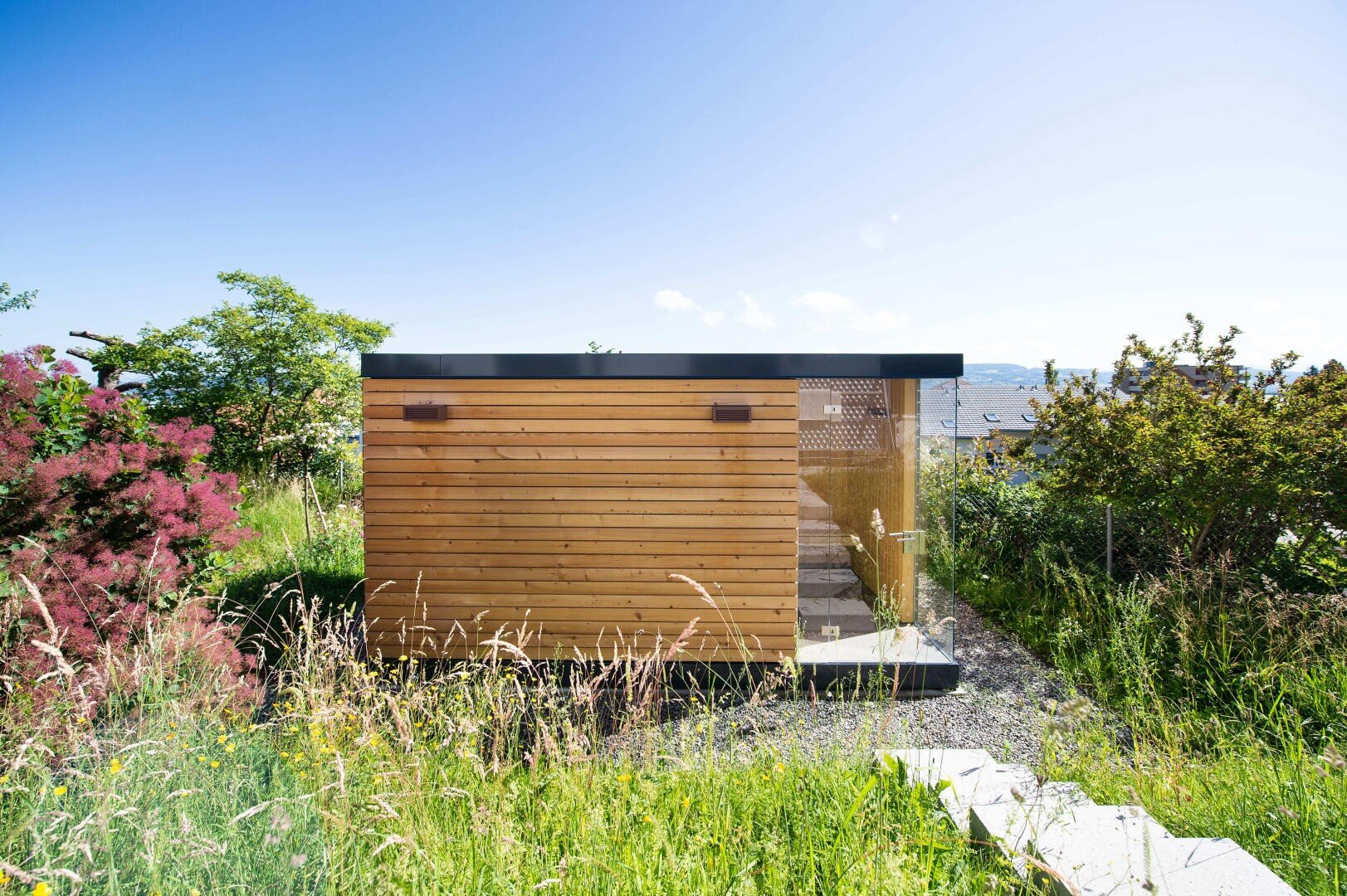 Ein kleiner moderner Holzschuppen mit Glasfront, umgeben von hohem Gras und Grün, unter einem klaren blauen Himmel. Ein Steinweg führt zum Eingang.