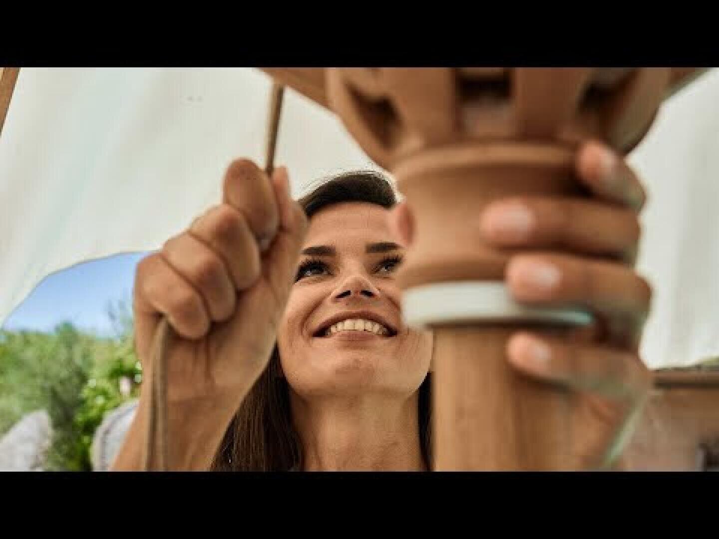 A woman smiles while working with her hands on a terracotta object outdoors, focusing intently on her task.