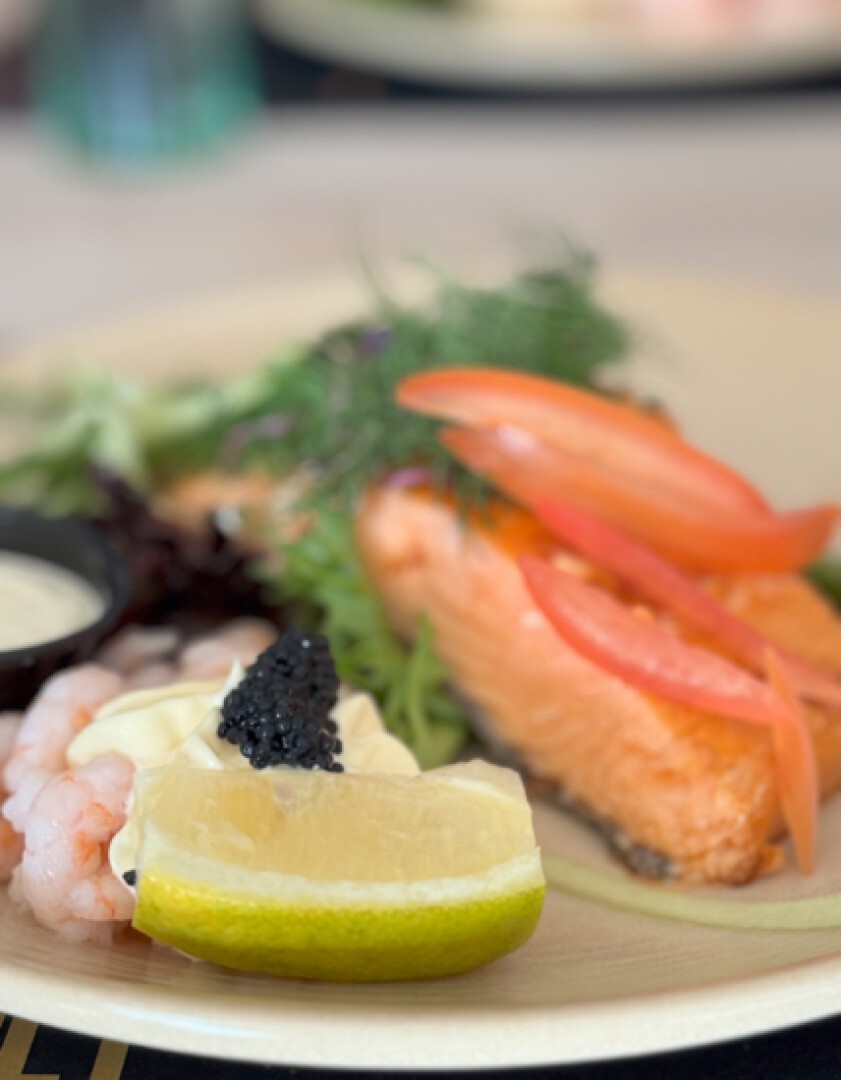 Eine Nahaufnahme eines Tellergerichts mit einem Lachsfilet, belegt mit Tomatenscheiben, Garnelen, einer Limettenspalte mit Kaviar, Gemüse und einer Beilage Sahnesauce.