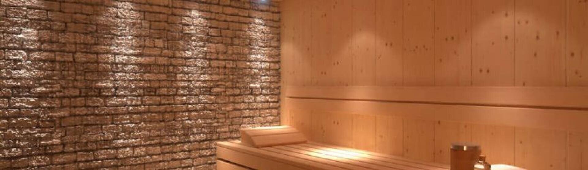 A modern sauna room with wooden benches and walls, featuring a textured stone accent wall illuminated by soft, recessed lighting. A small wooden bucket and ladle are placed on a bench.