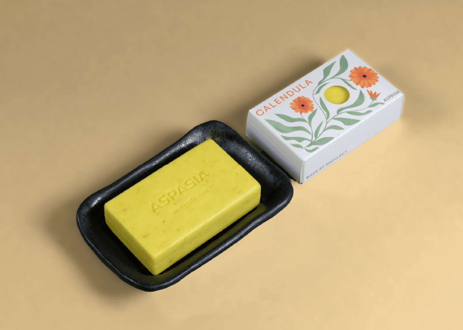 A yellow bar of soap with the inscription ASPASIA lies in a black soap dish next to a white box with orange flowers and green leaves bearing the inscription CALENDULA.